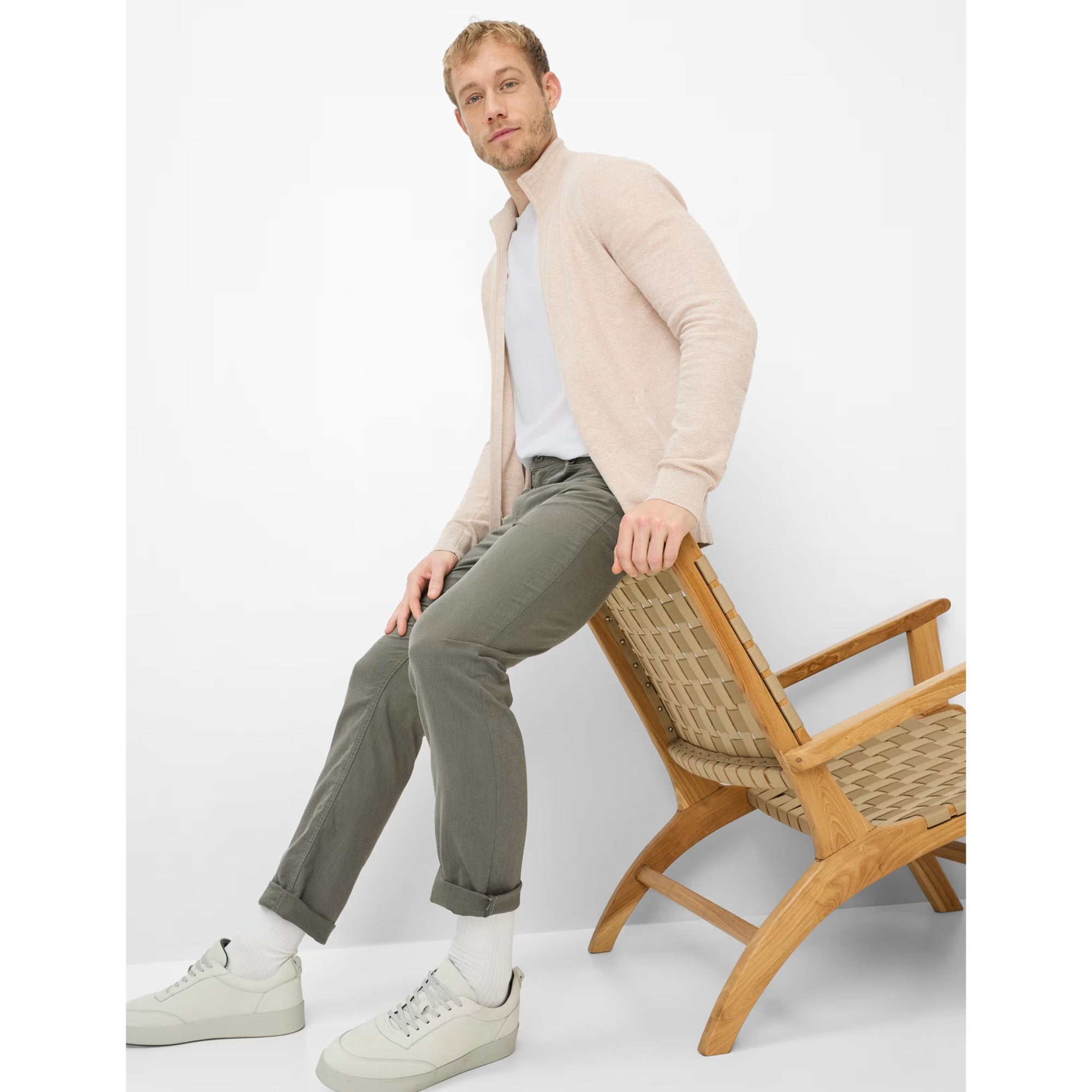 Man sitting on a wooden chair wearing a beige cardigan, white shirt, green pants, and white sneakers.