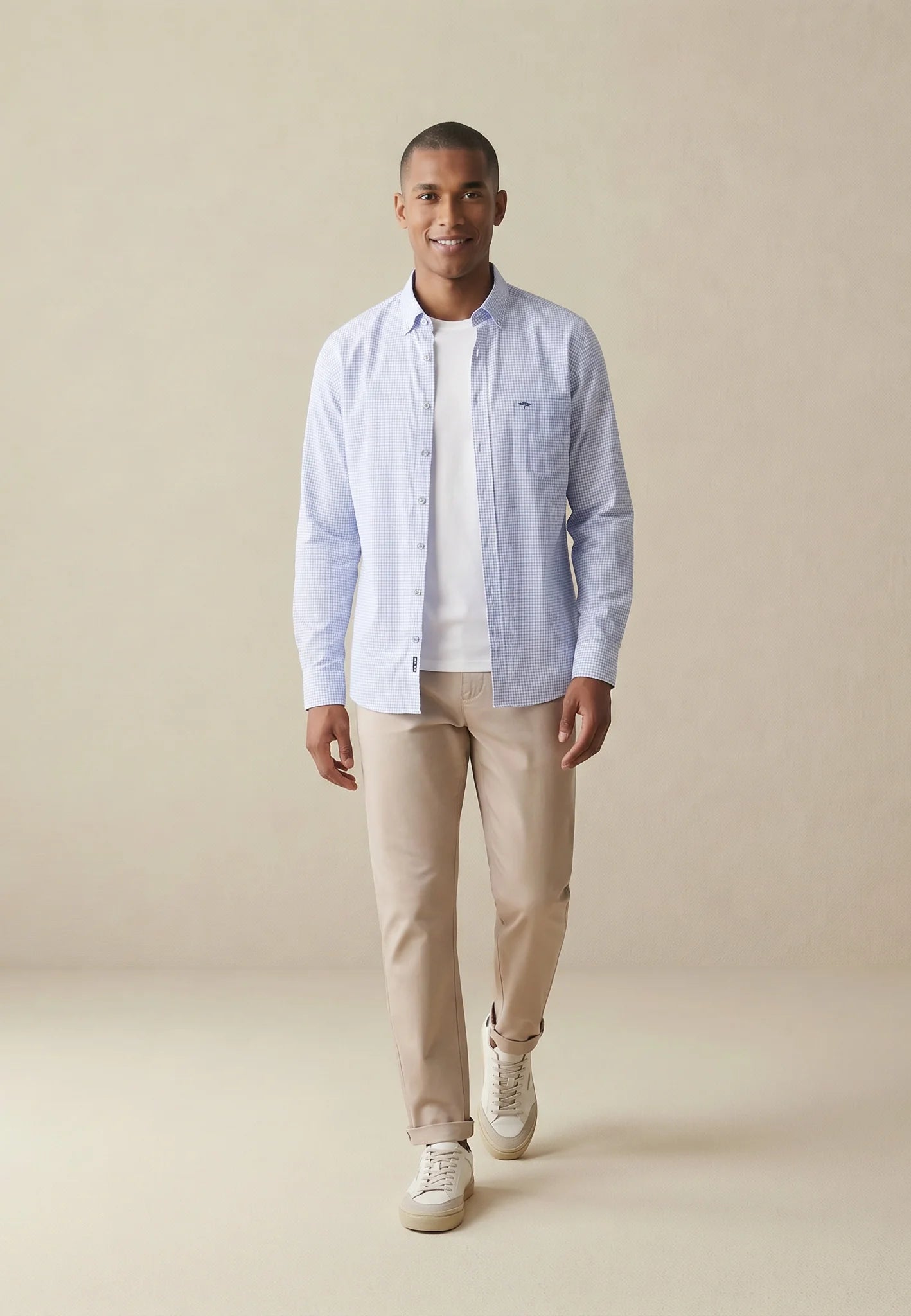 Man wearing a light blue shirt, white t-shirt, beige pants, and white sneakers on a beige background