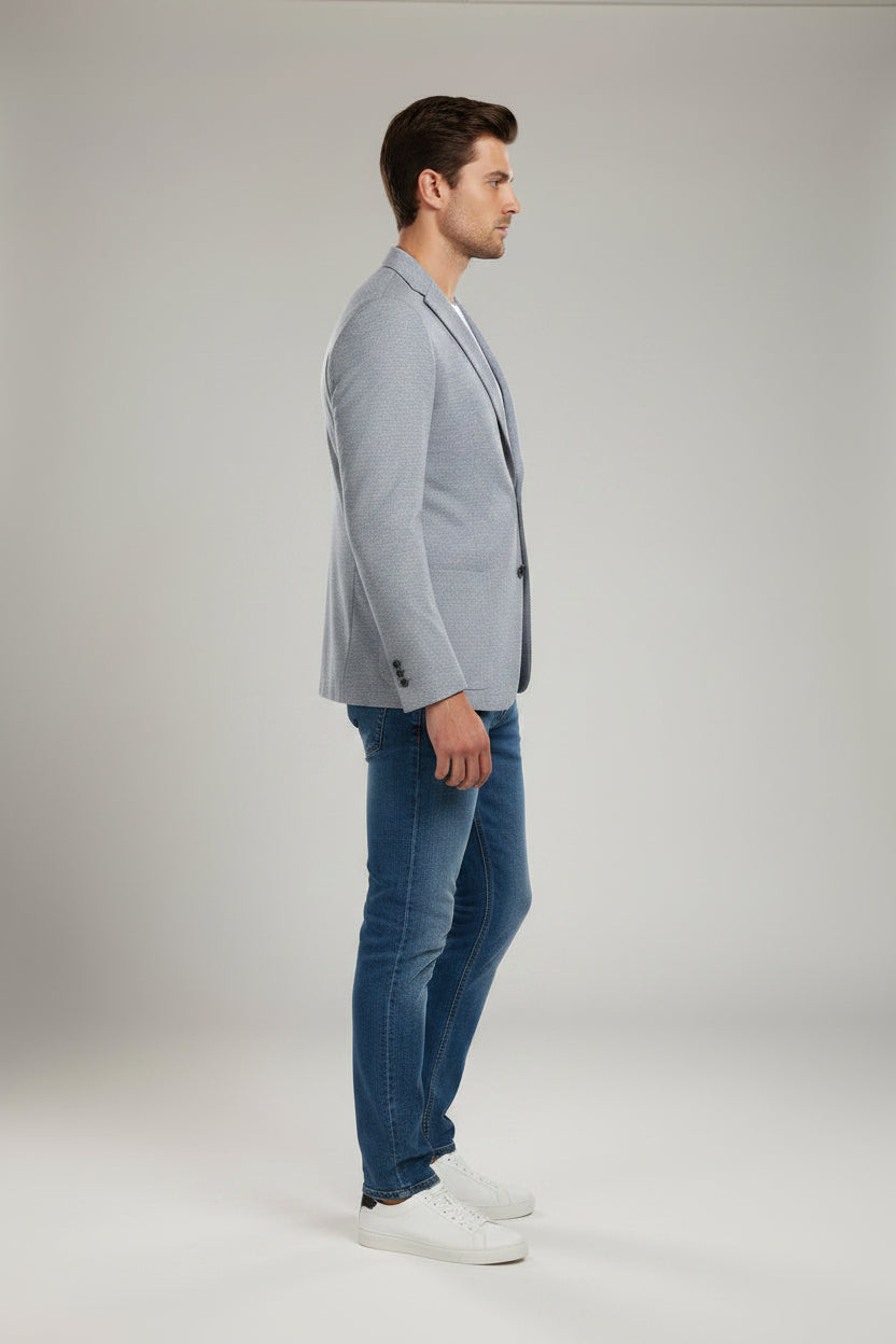 A man modeling a light blue textured blazer over a white t-shirt, blue jeans, and white sneakers, standing against a neutral light gray studio background.