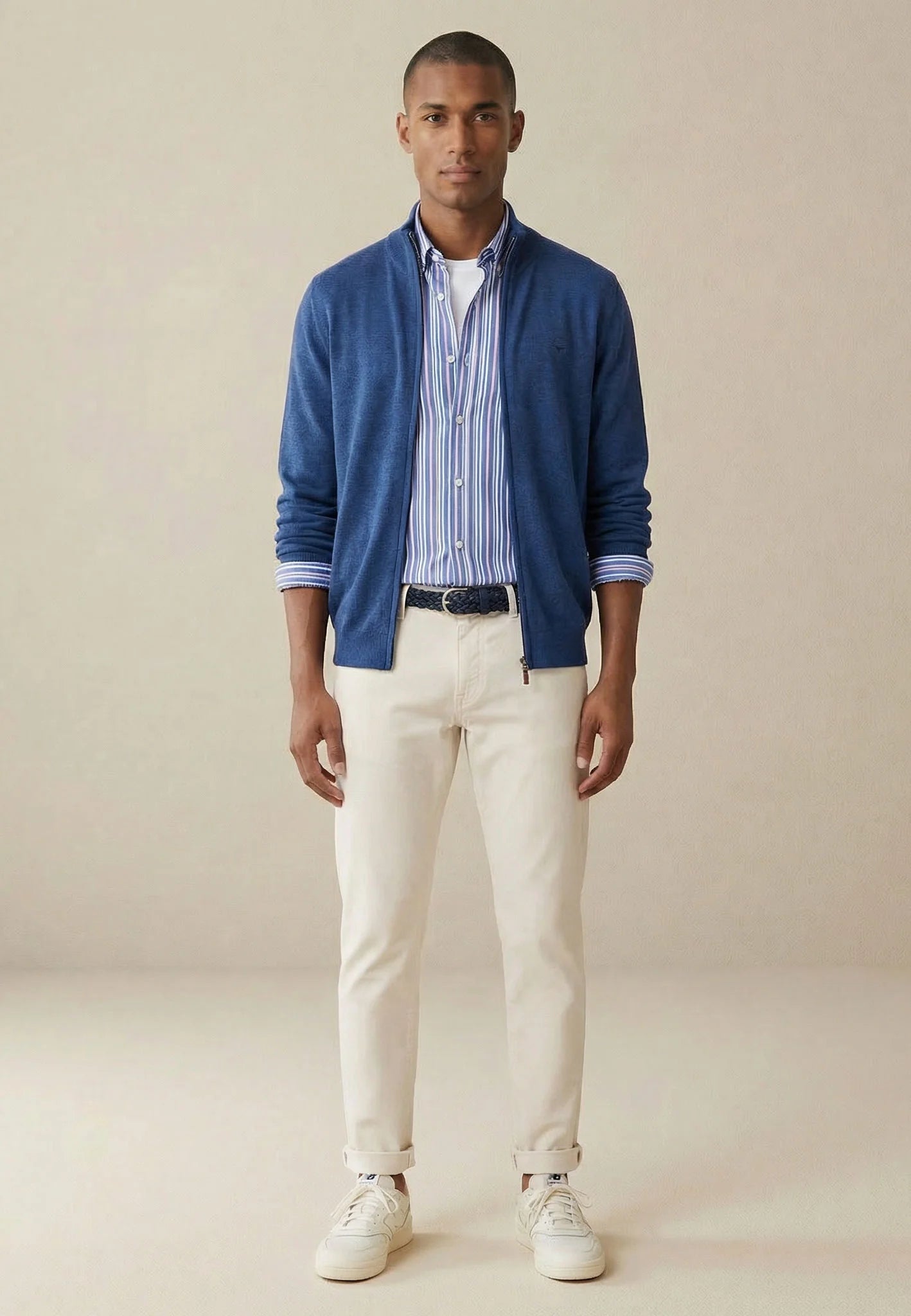 Man wearing a blue cardigan, striped shirt, white pants, and sneakers on a beige background