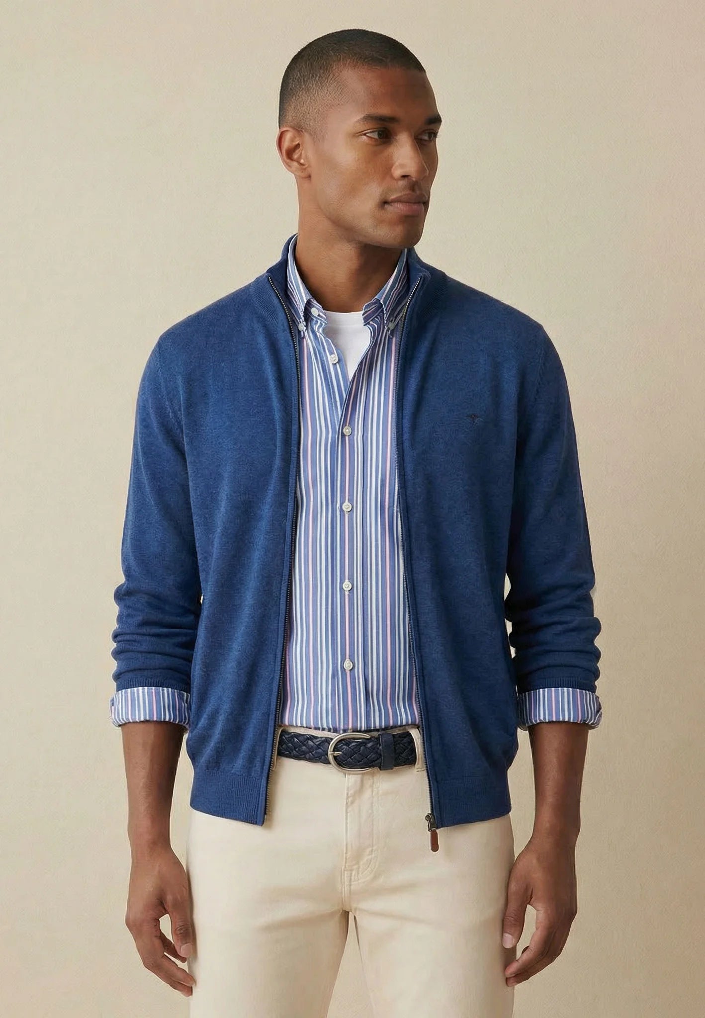 Man wearing a blue cardigan over a striped shirt with beige pants on a beige background