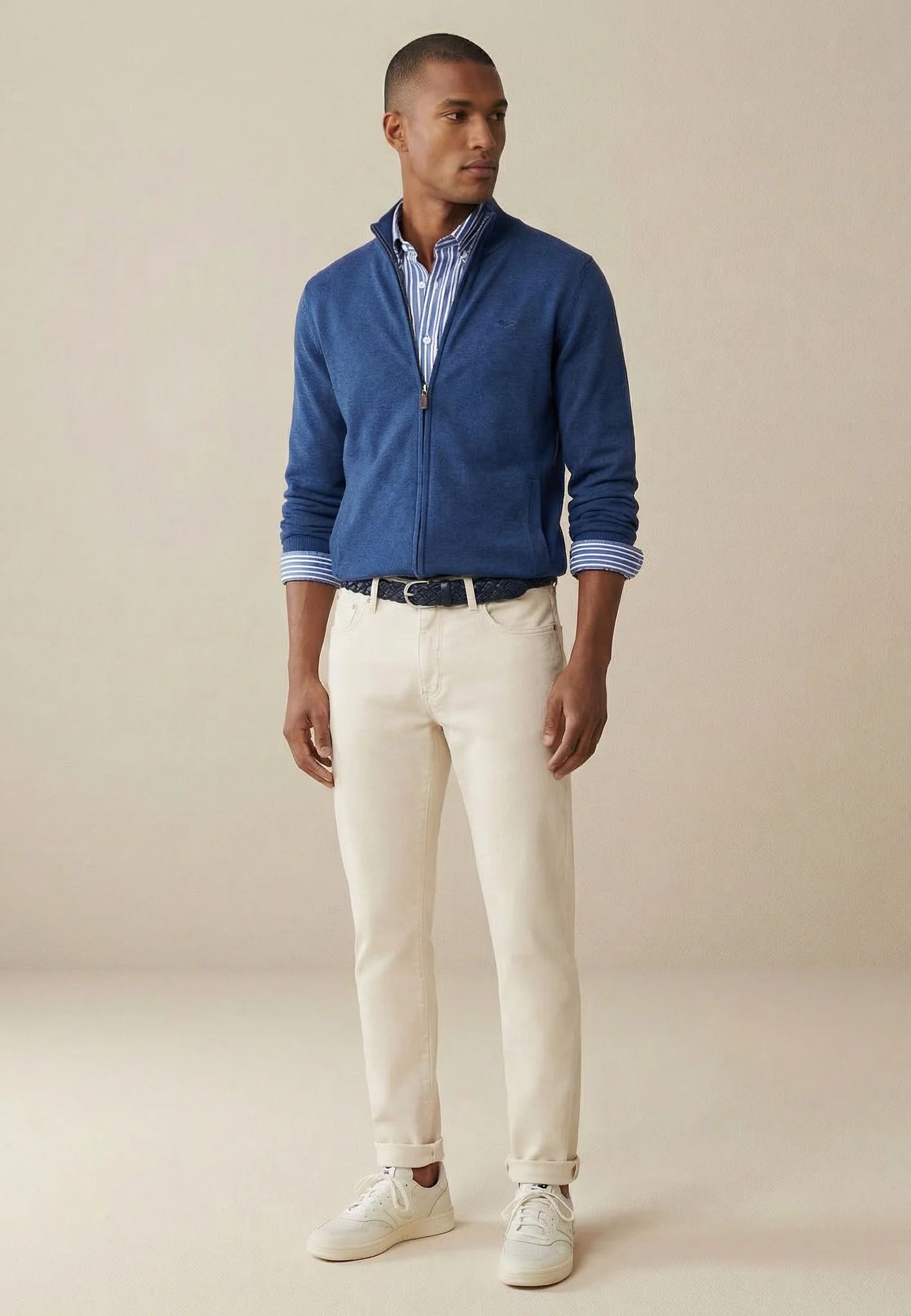 Man wearing a blue jacket, checkered shirt, and white pants on a beige background