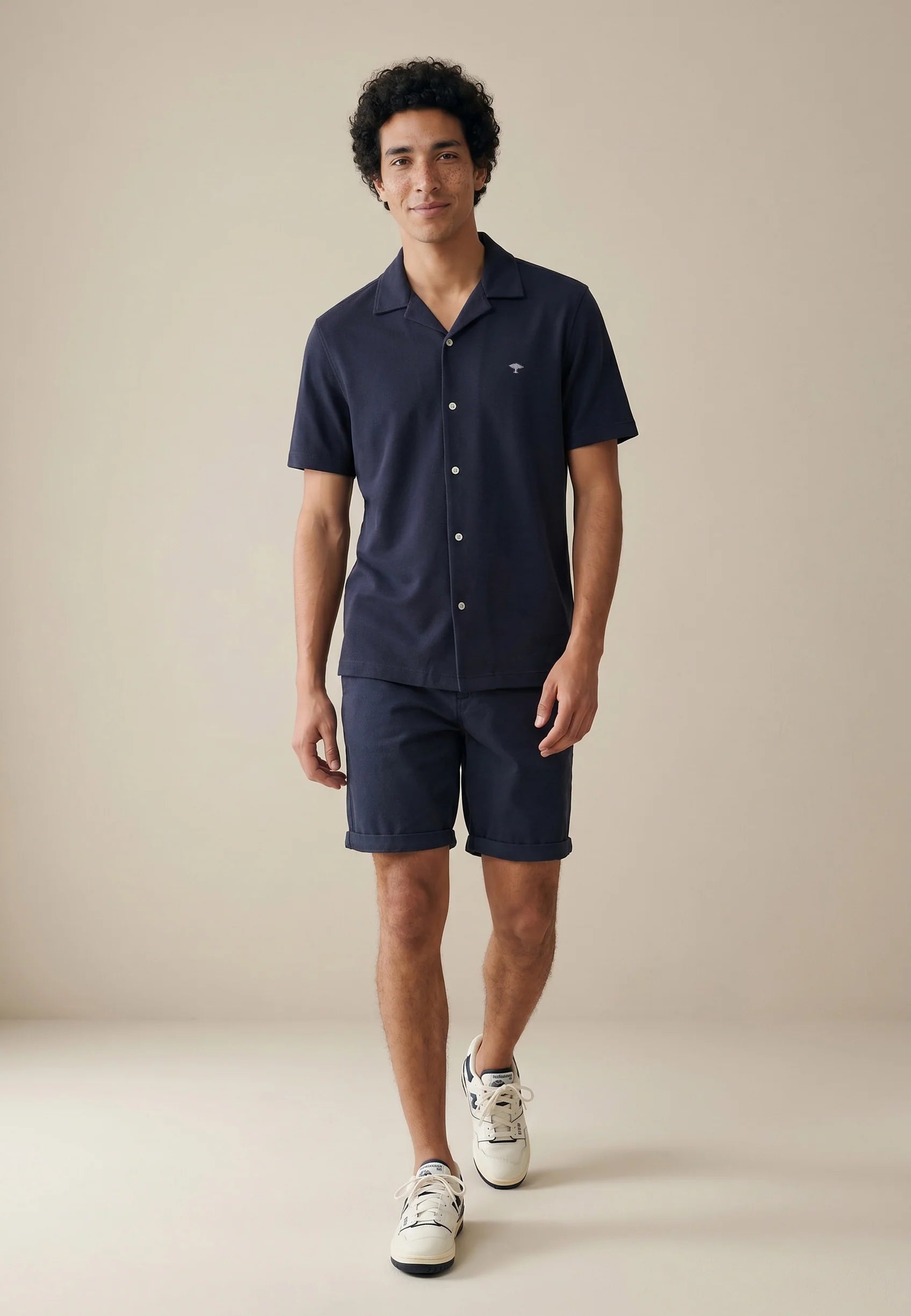 Man wearing a navy shirt and shorts with white sneakers on a beige background