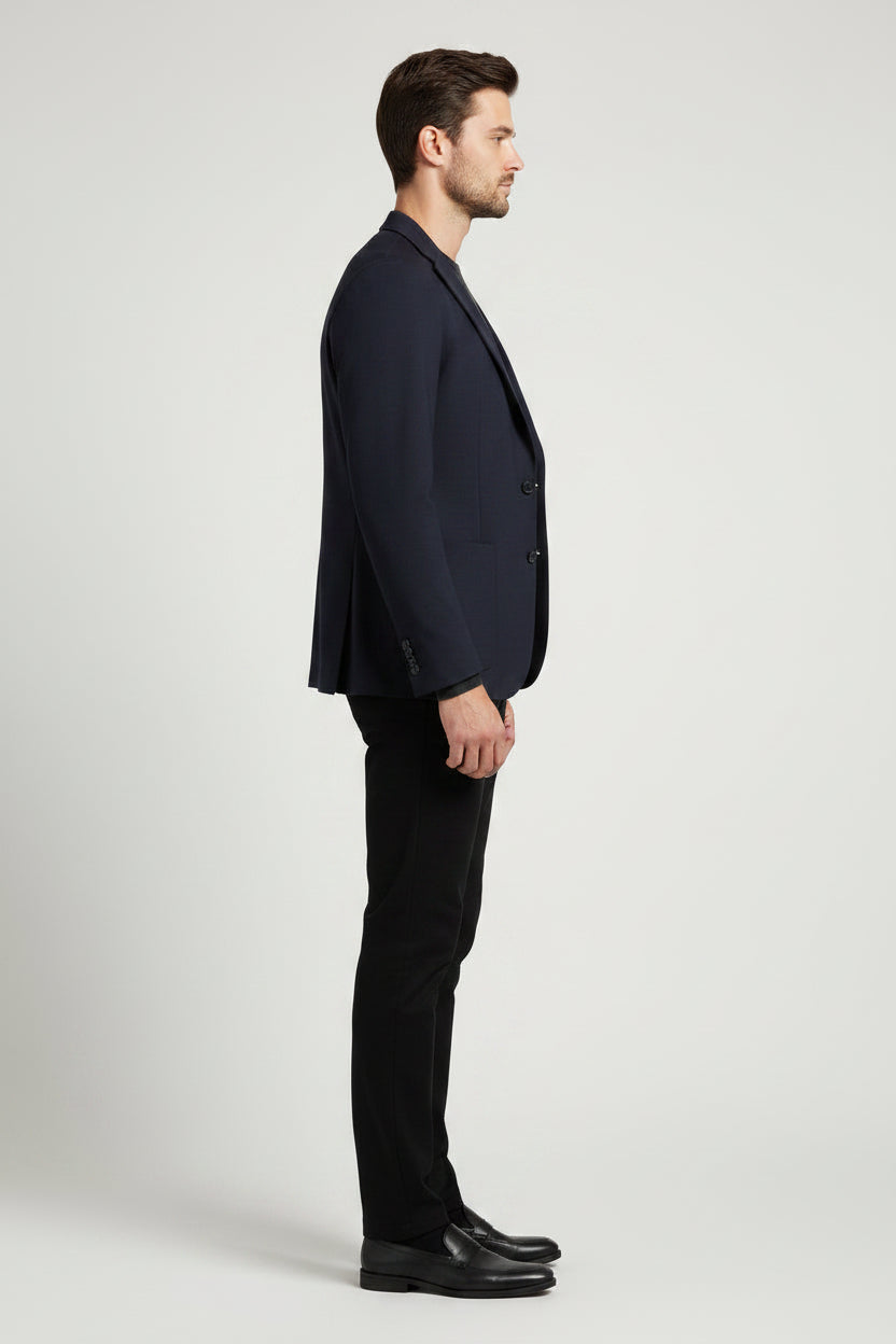 A man modeling a dark navy blue blazer, black crewneck sweater, black trousers, and black dress shoes against a light gray studio background.