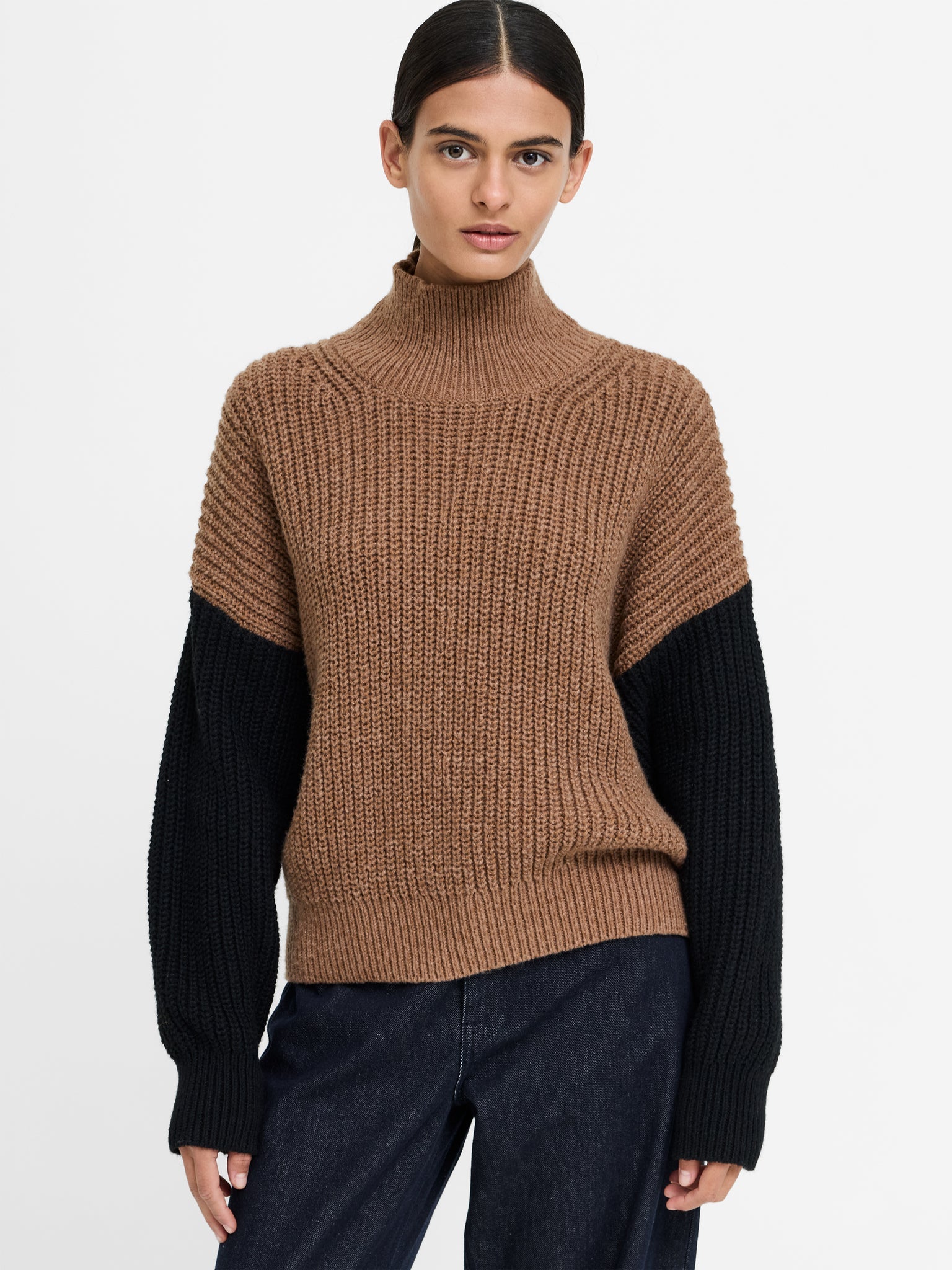 A woman modeling a ribbed knit turtleneck sweater with a brown body and black sleeves, paired with dark blue jeans, against a plain light gray background.