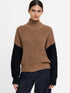 A woman modeling a ribbed knit turtleneck sweater with a brown body and black sleeves, paired with dark blue jeans, against a plain light gray background.