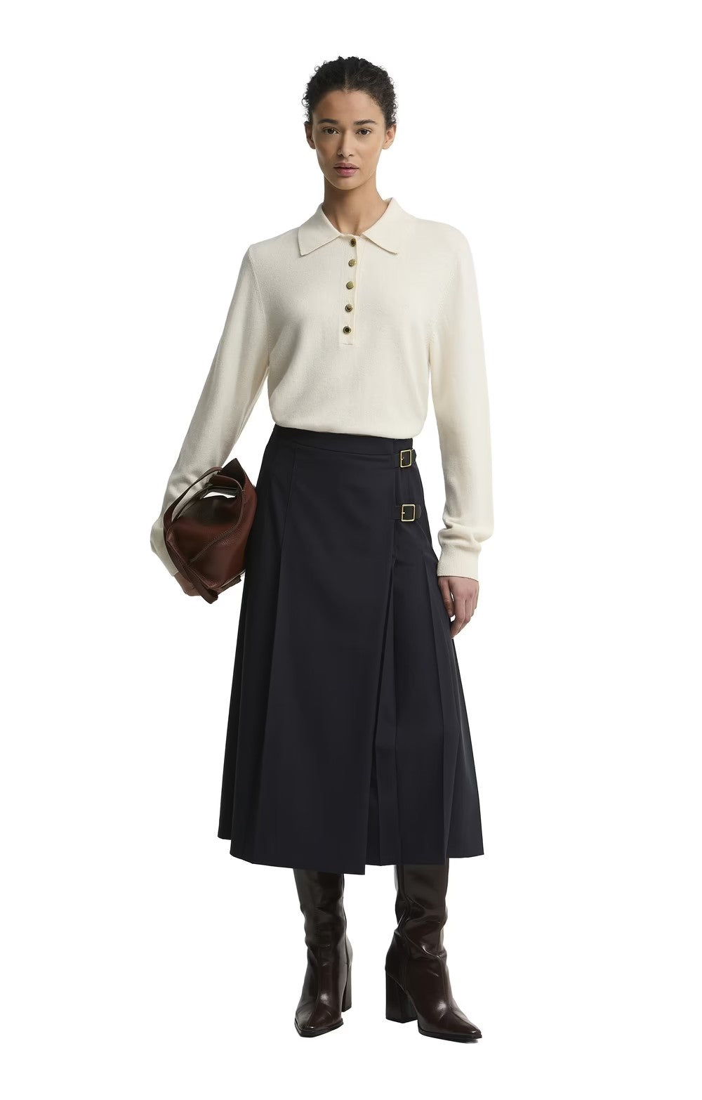 A model stands against a white studio background wearing a cream long-sleeve polo-style top with three front buttons, a dark navy midi skirt with belt details, and dark brown knee-high boots. She holds a brown leather bag.