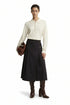 A model stands against a white studio background wearing a cream long-sleeve polo-style top with three front buttons, a dark navy midi skirt with belt details, and dark brown knee-high boots. She holds a brown leather bag.