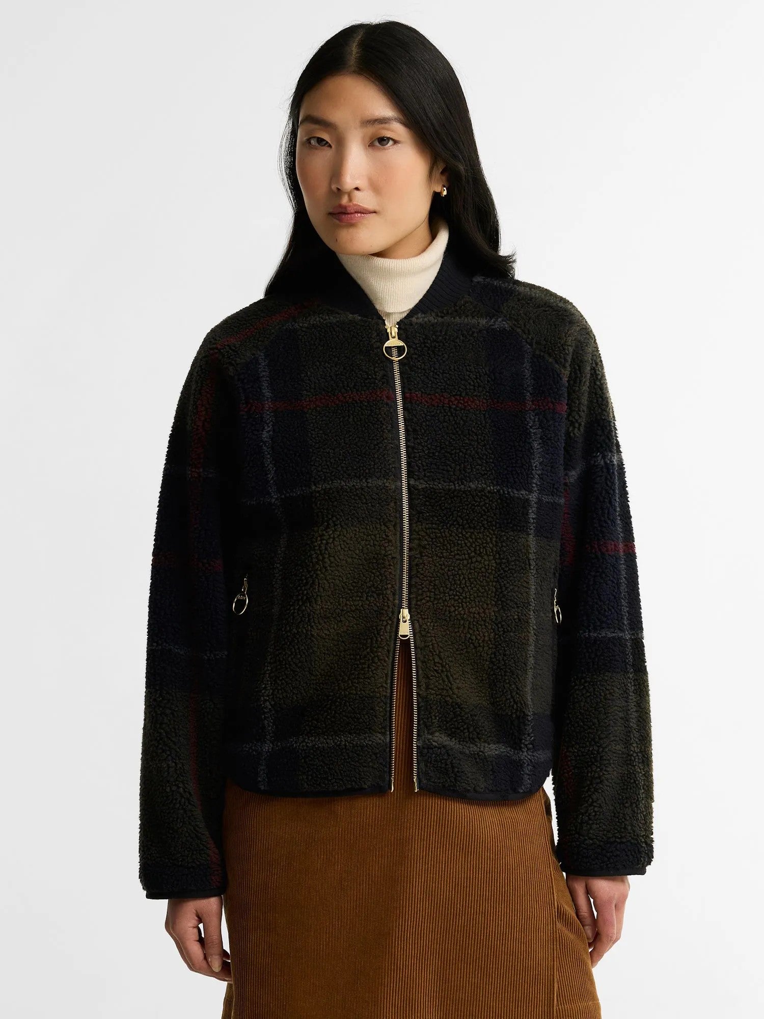A woman wearing a cropped zip-front Barbour flecce jacket with a dark green and black plaid pattern, styled with a white turtleneck and brown skirt against a light gray studio background.