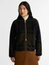 A woman wearing a cropped zip-front Barbour flecce jacket with a dark green and black plaid pattern, styled with a white turtleneck and brown skirt against a light gray studio background.