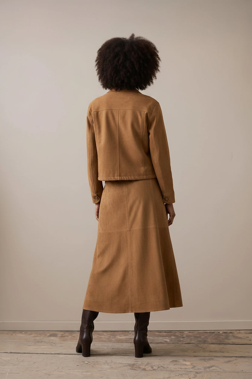 A model stands in a studio wearing a camel brown suede jacket and matching midi skirt set with a white collared shirt and dark brown ankle boots, against a neutral beige background.