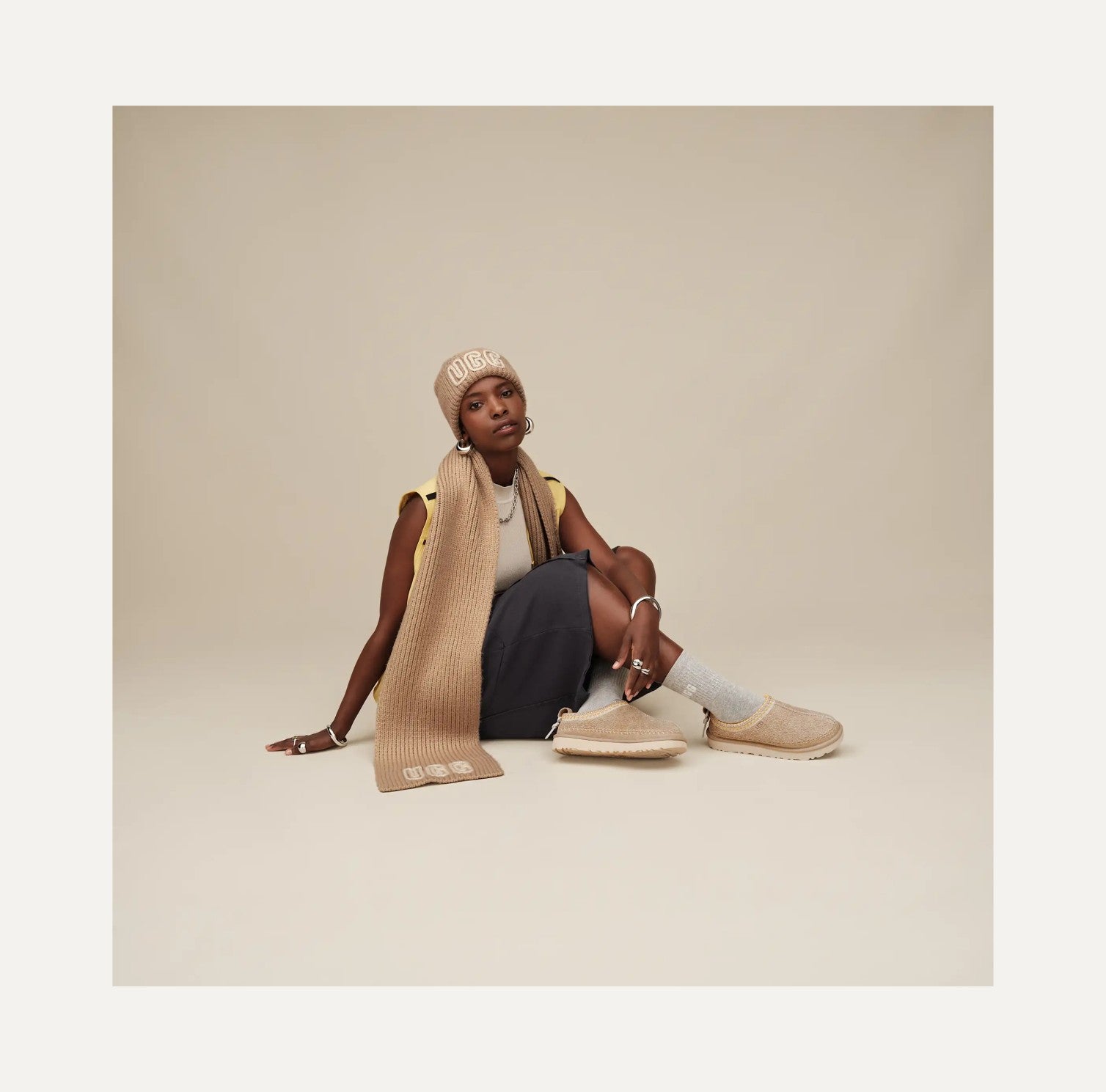 Woman sitting on the floor wearing a beige knit sweater, pants, and shoes against a plain background