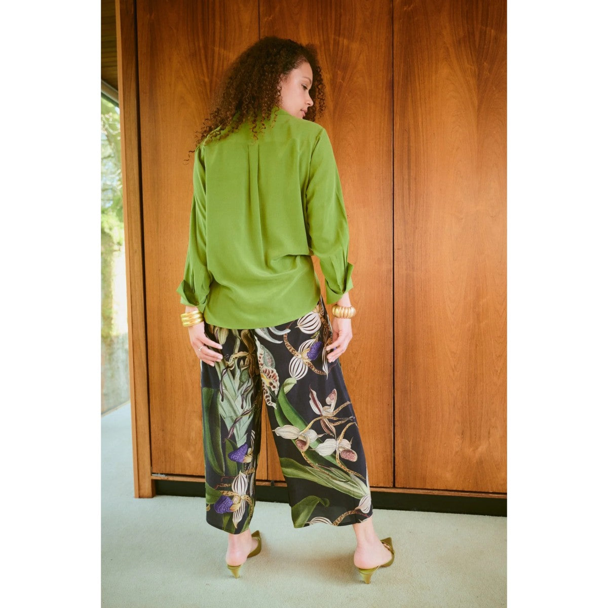 Woman wearing a green blouse and floral pants standing in front of a wooden door.