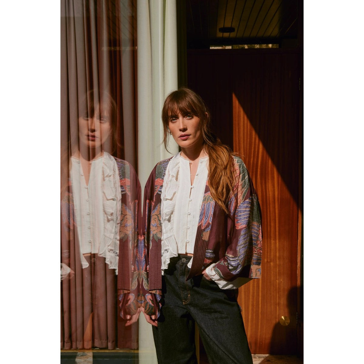 Woman wearing a patterned cardigan and scarf in a room with curtains.