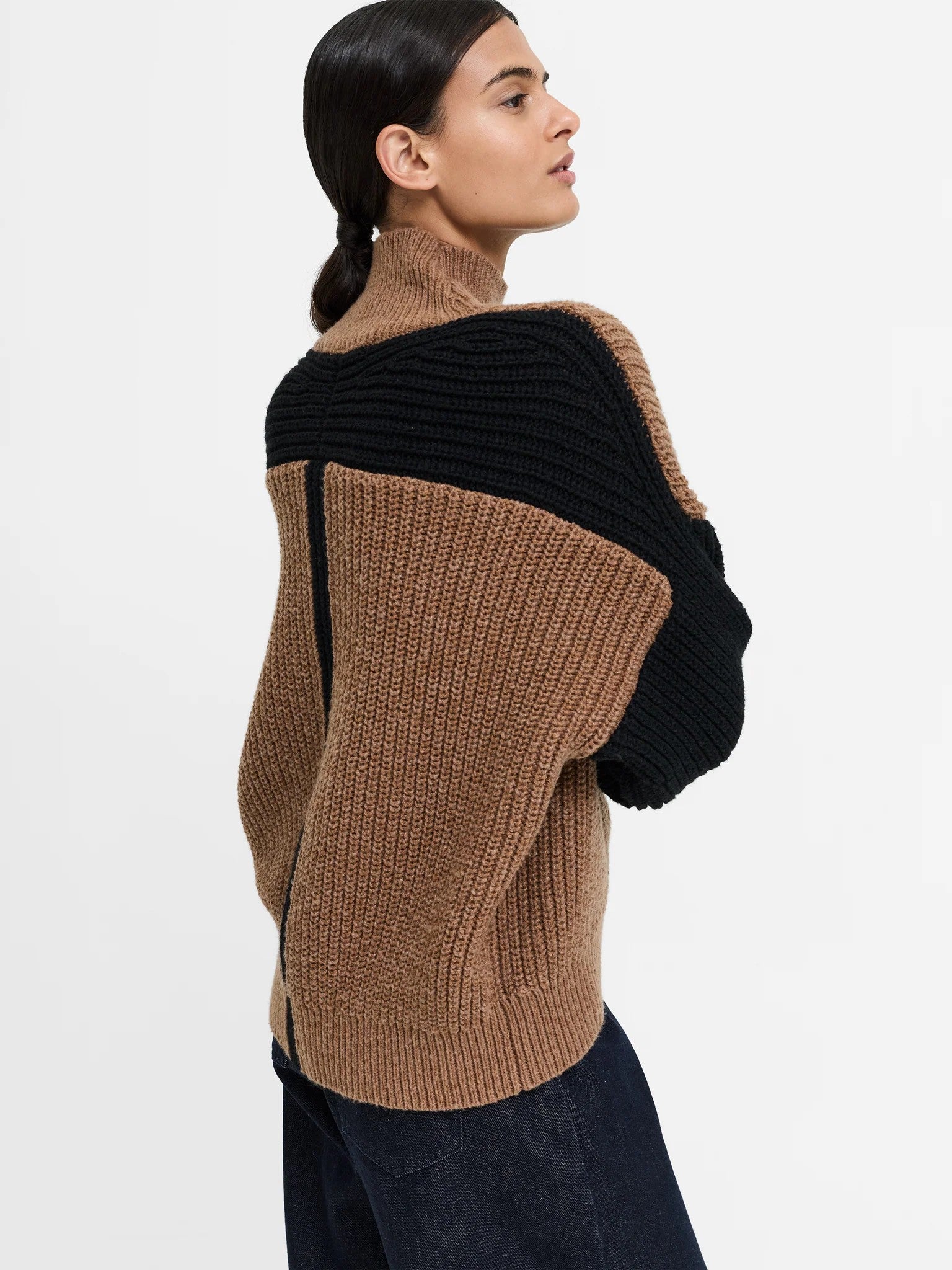 A woman wearing a chunky ribbed knit sweater with a color-blocked design in brown and black, shown from a side-back angle against a plain light gray studio background.