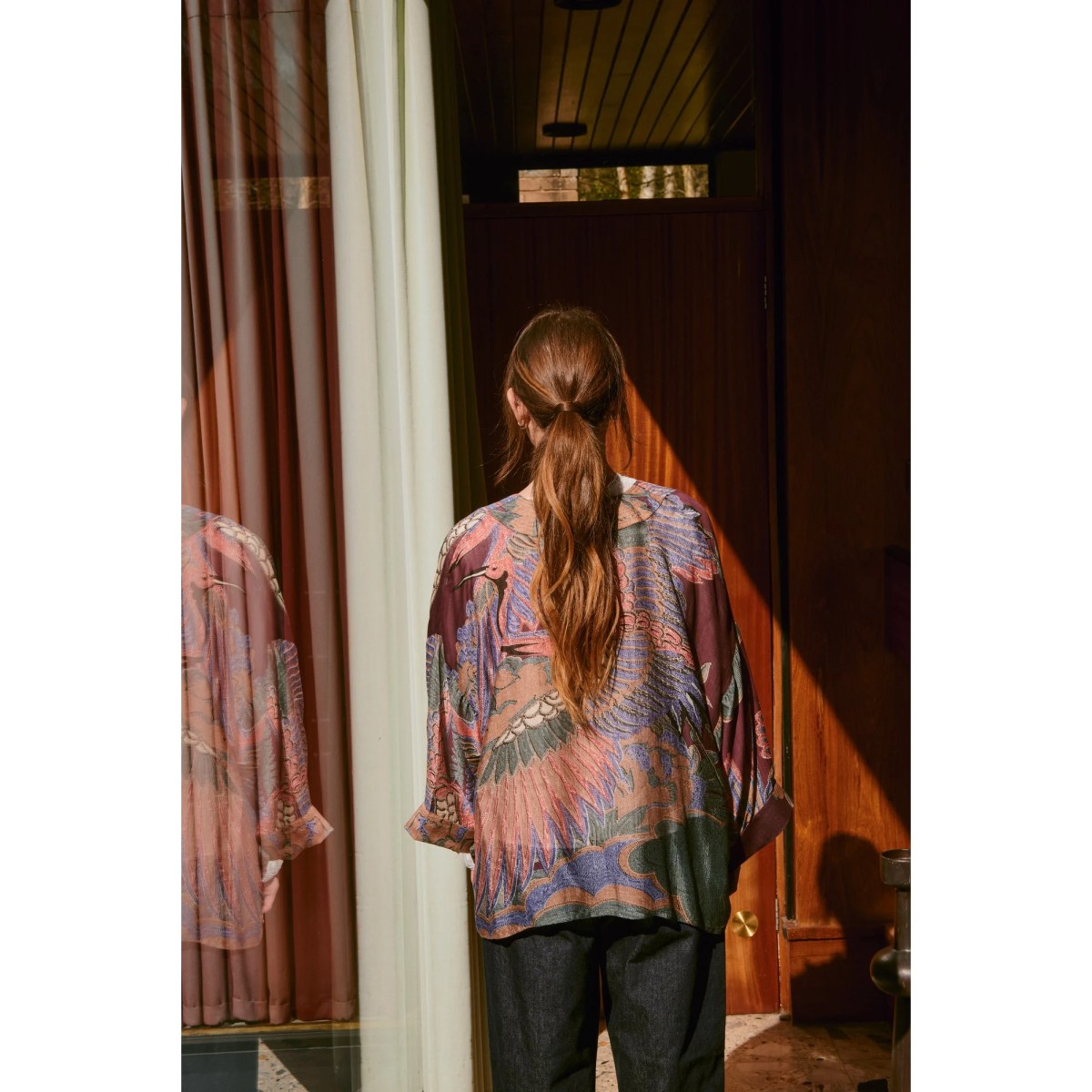 Person wearing a patterned shirt standing in a room with wooden walls and a mirror.