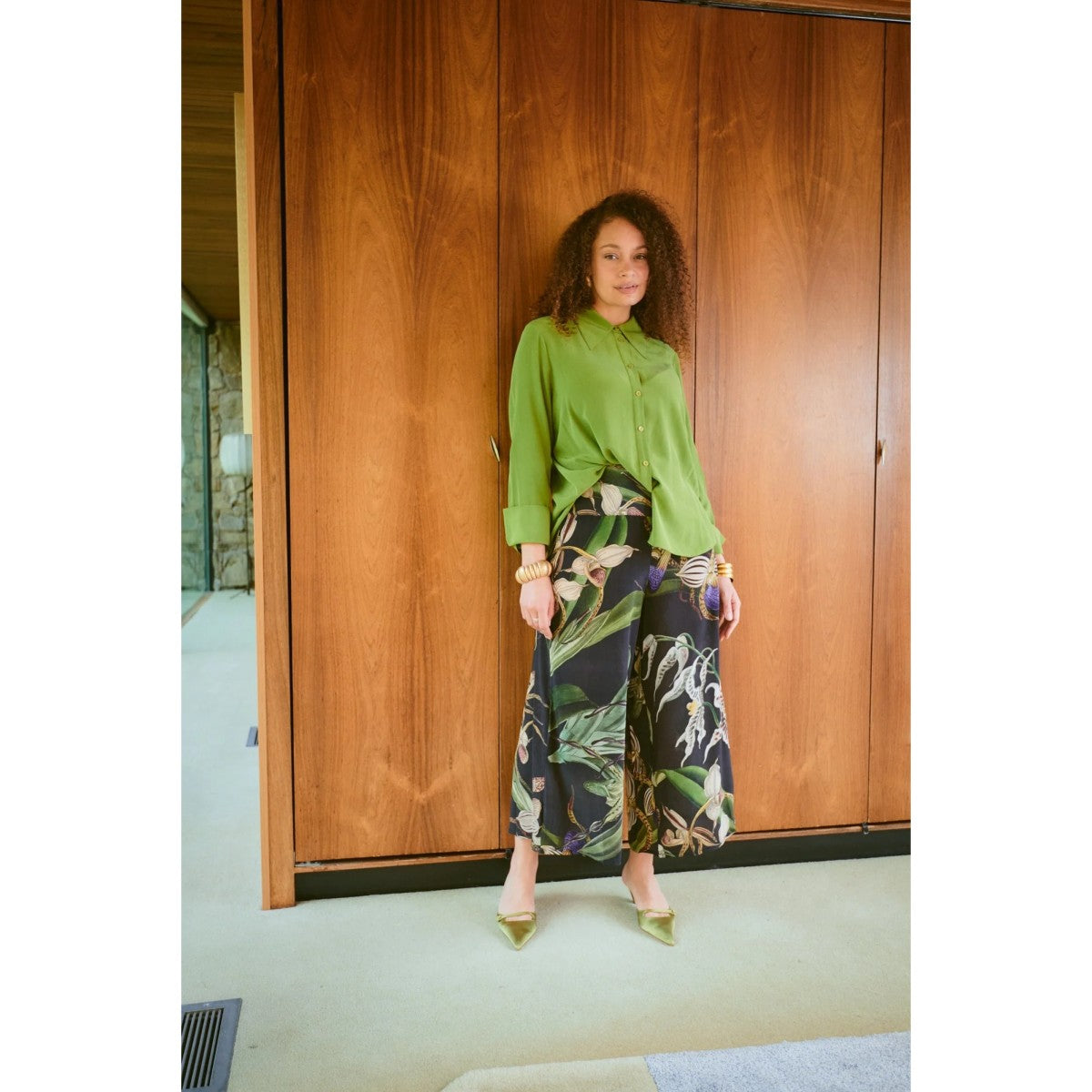 Woman in a green top and floral pants standing in front of wooden doors.