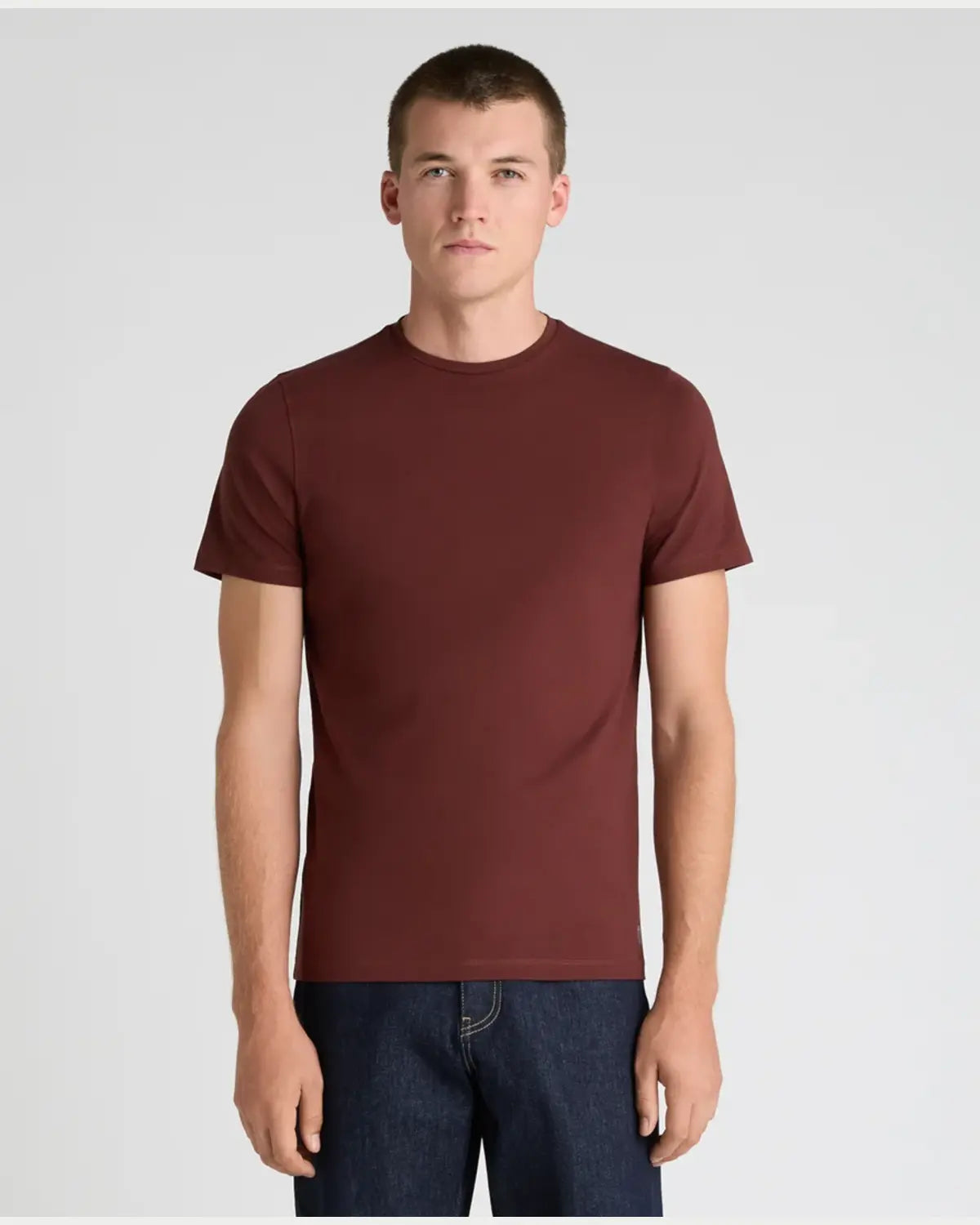 A man wearing a short-sleeve crewneck t-shirt in a deep burgundy color, paired with dark blue jeans, standing against a light gray studio background.