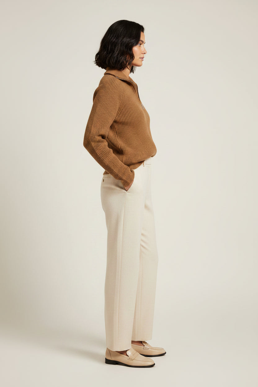 A woman stands against a plain light beige studio background wearing a camel brown ribbed knit zip-up sweater, cream-colored wide-leg trousers, and beige loafers.