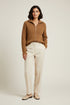 A woman stands against a plain light beige studio background wearing a camel brown ribbed knit zip-up sweater, cream-colored wide-leg trousers, and beige loafers.