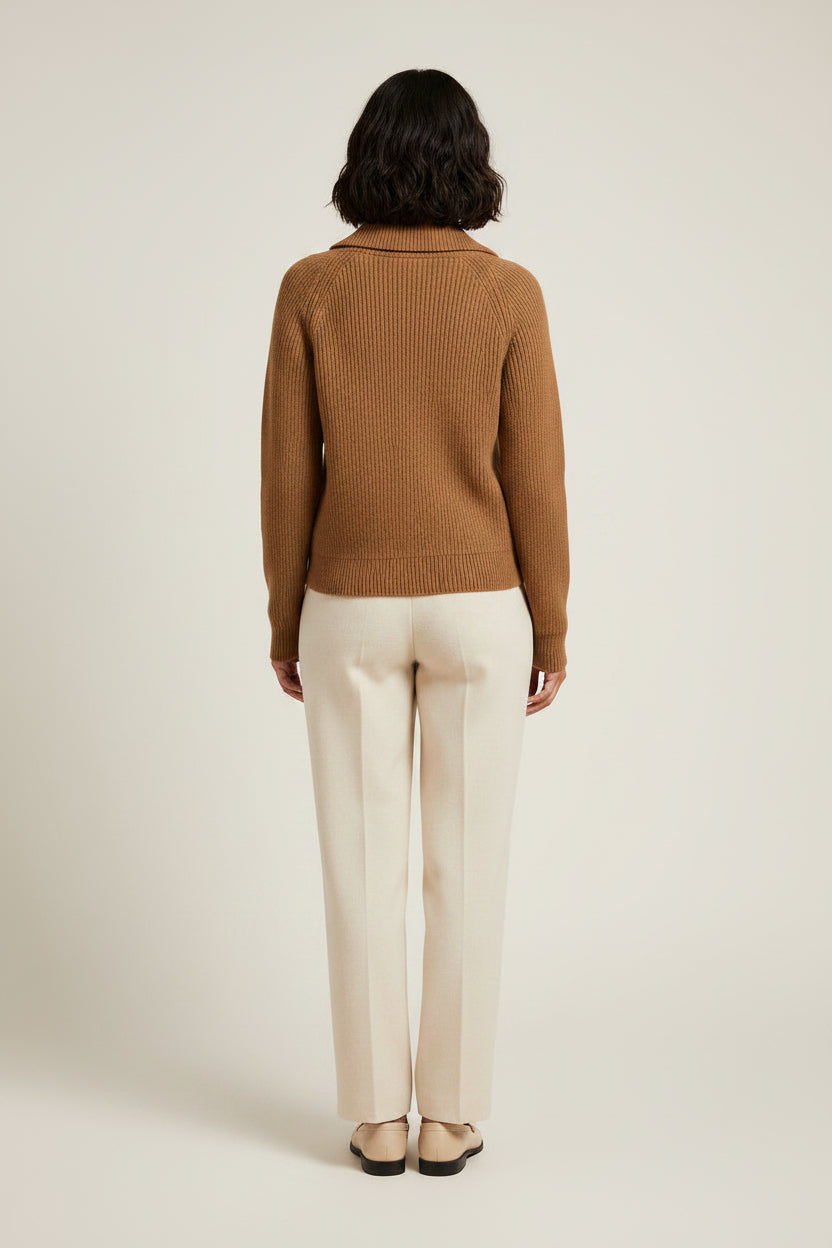 A woman stands against a plain light beige studio background wearing a camel brown ribbed knit zip-up sweater, cream-colored wide-leg trousers, and beige loafers.