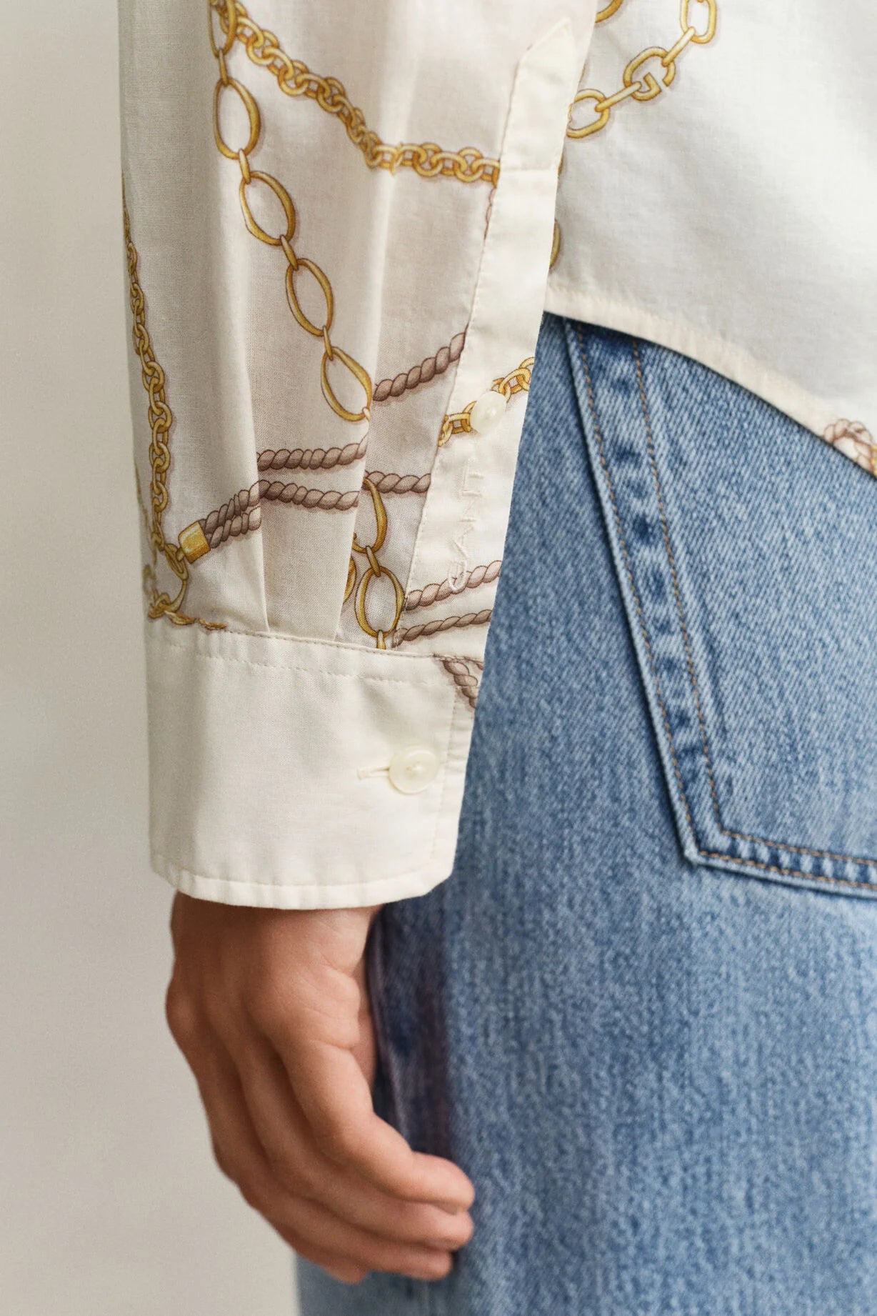 Close-up of a person wearing a white blouse with gold chain pattern and blue jeans.