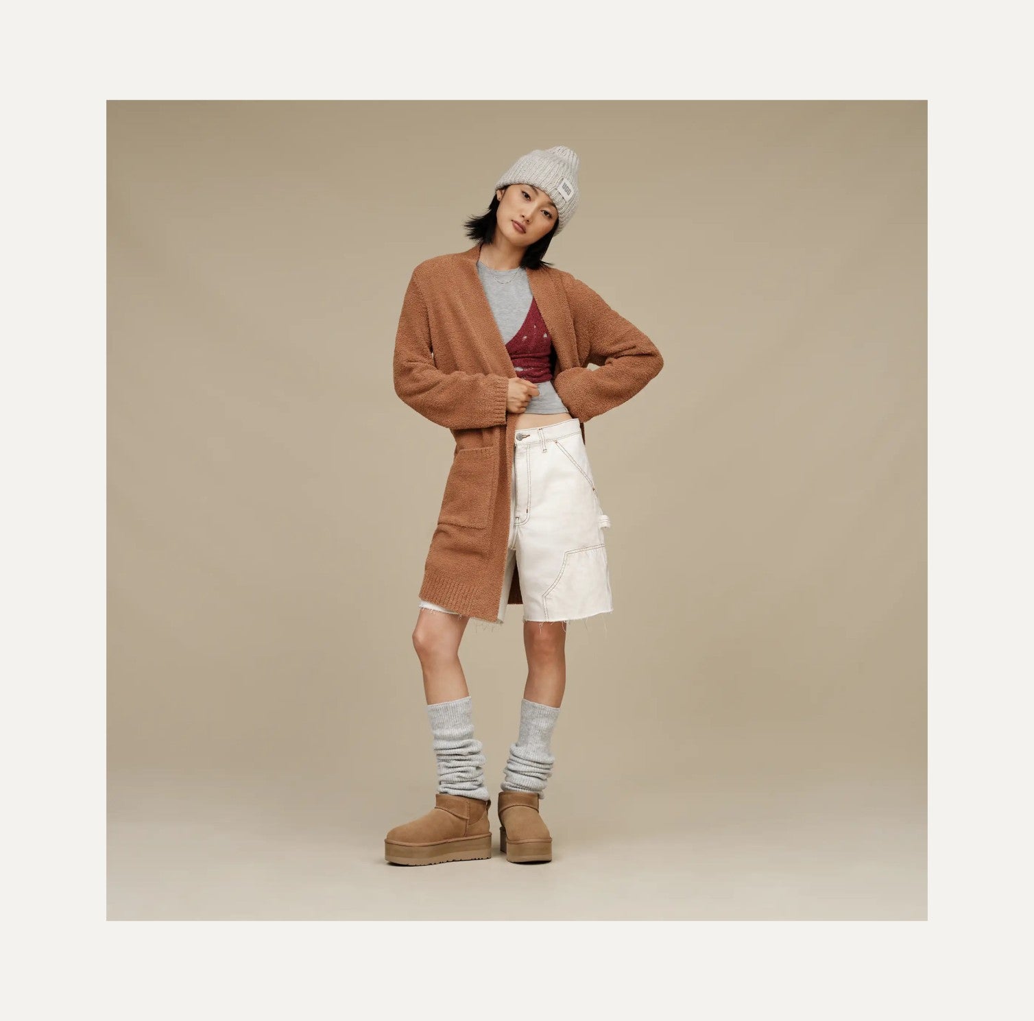 Person wearing a brown cardigan, white shorts, and beige boots on a beige background