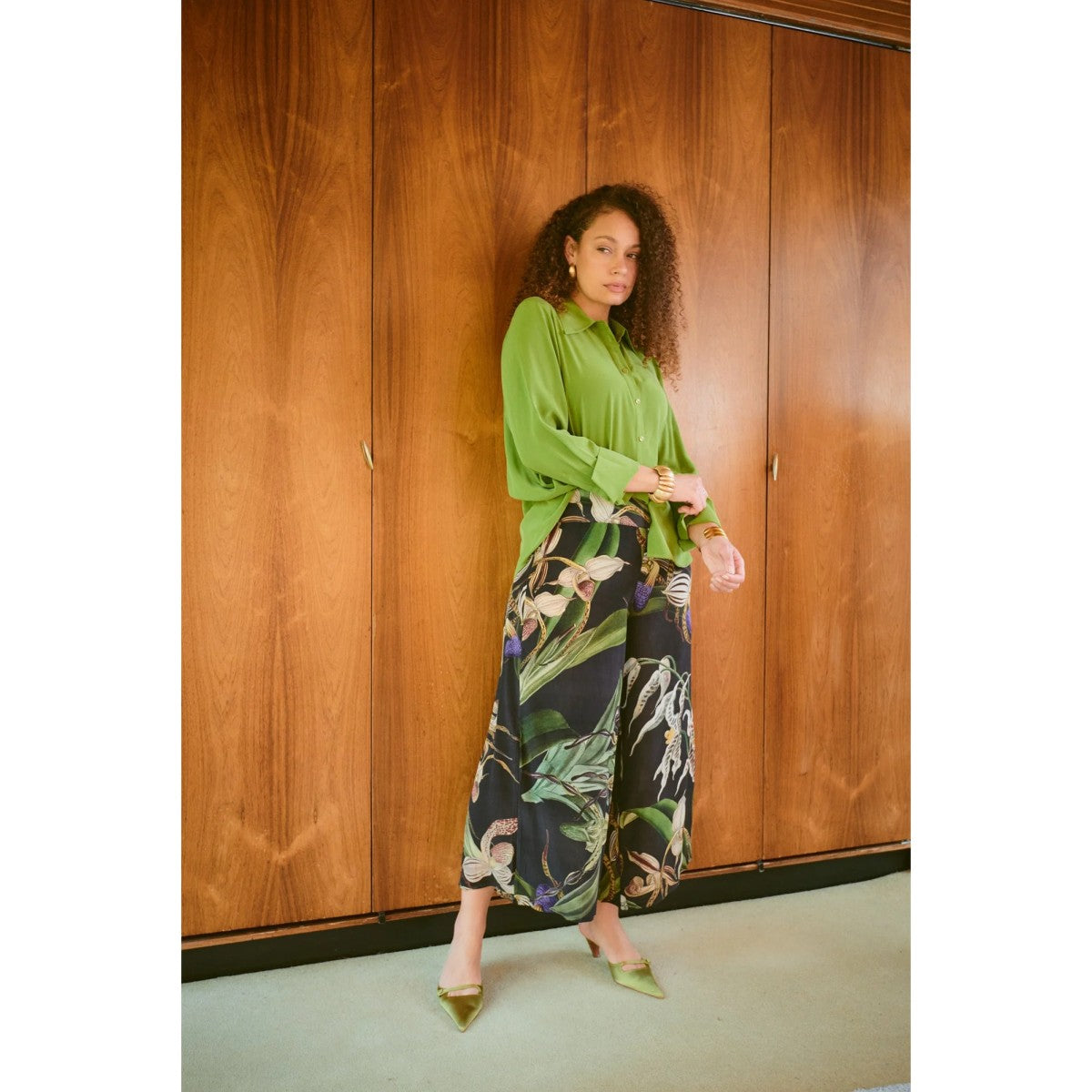 Woman wearing a green blouse and patterned pants standing against a wooden wall.