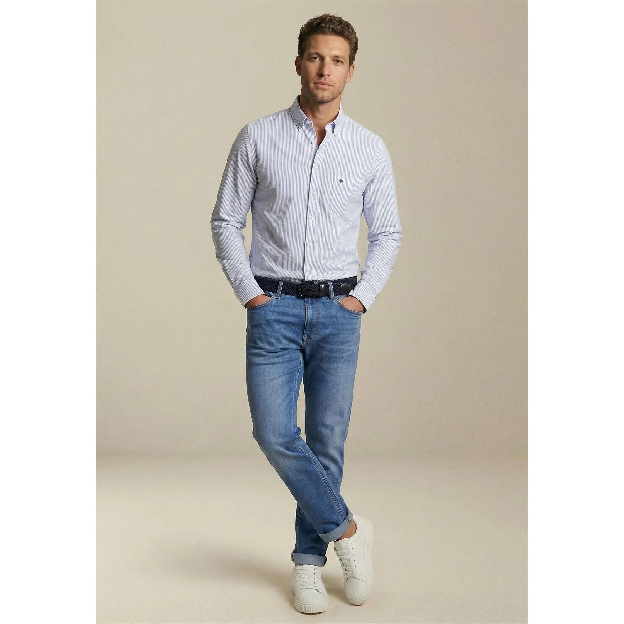 Man wearing a light blue shirt, jeans, and white sneakers against a beige background