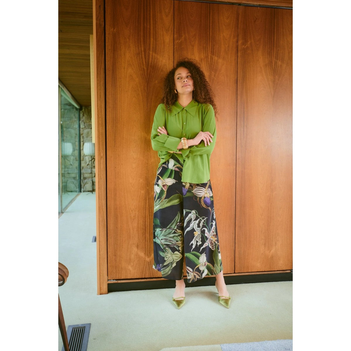 Woman in a green top and floral pants standing in a room with wooden walls.