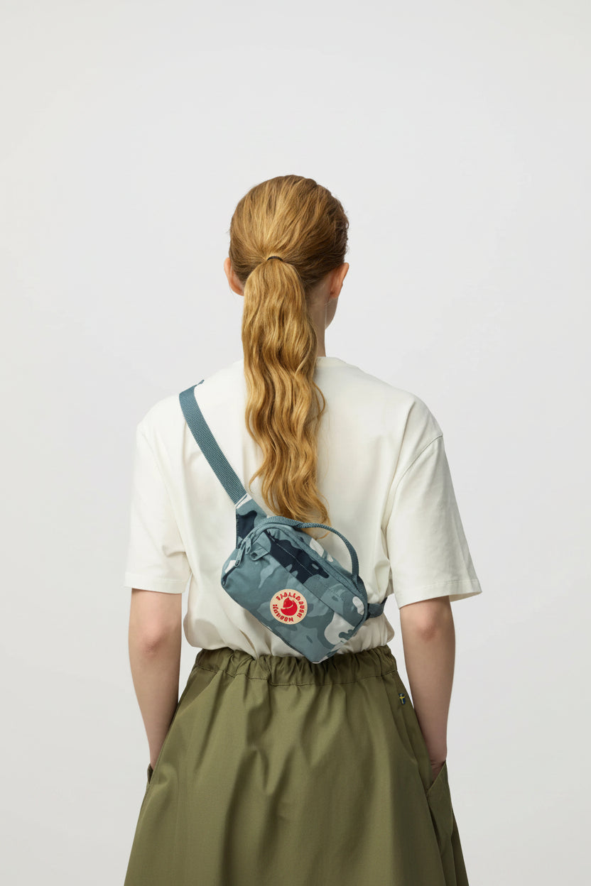 Person wearing a blue crossbody bag with a visible brand logo on a plain background
