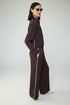A woman wearing a French Connection dark brown wide-leg tracksuit with white piping details, standing in profile against a split white and wood-paneled wall.