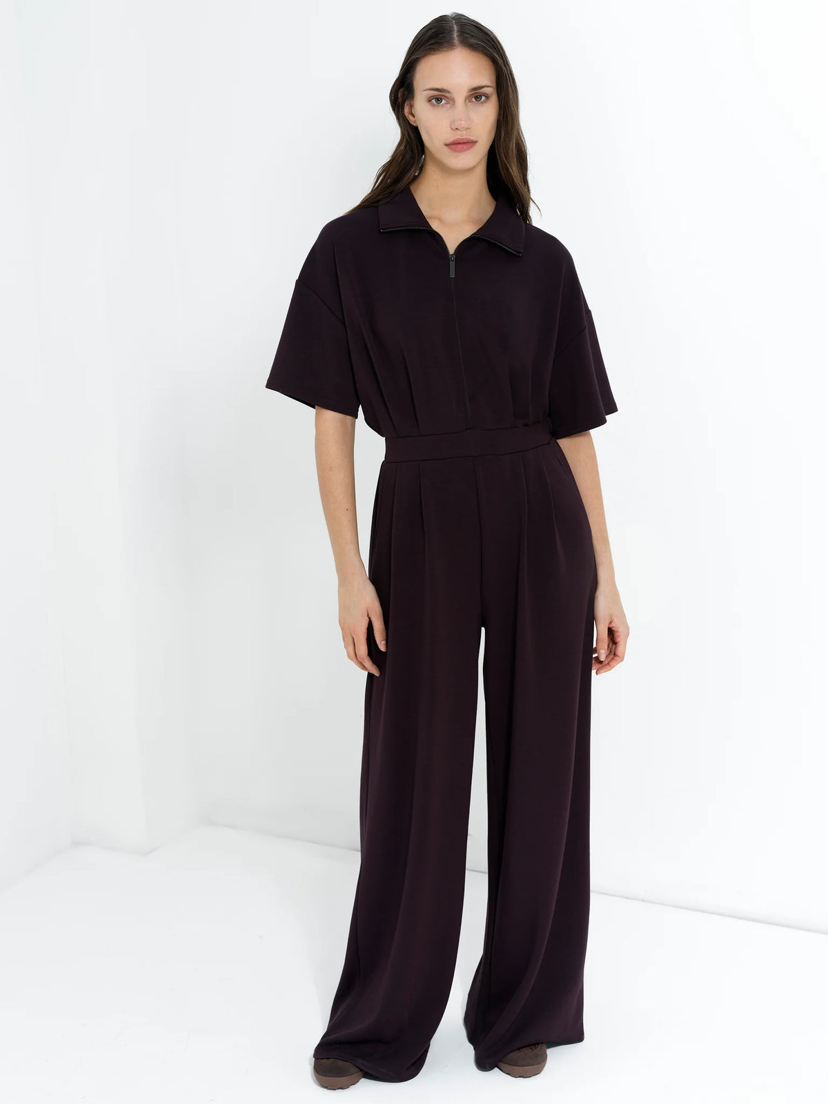 A woman modeling a French Connection  dark plum-colored short-sleeve jumpsuit with a half-zip collar and wide legs, standing against a clean white studio background.