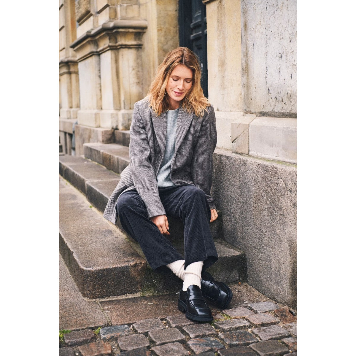 Woman sitting on stone steps wearing a gray coat and black shoes in an urban setting.