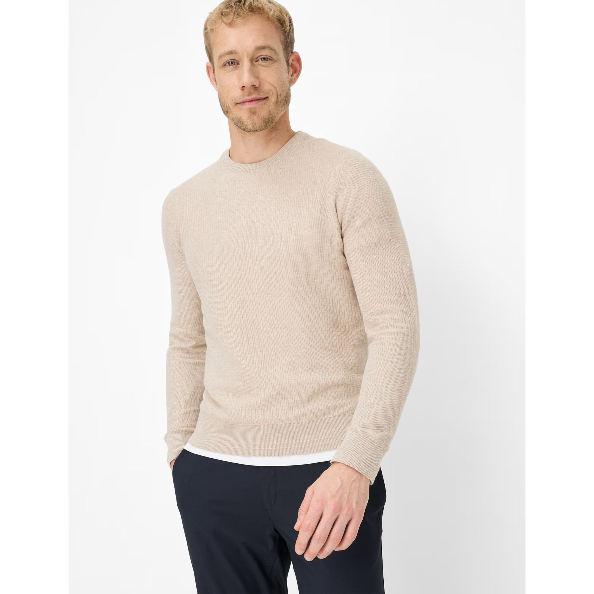 Man wearing a beige sweater and dark pants on a white background