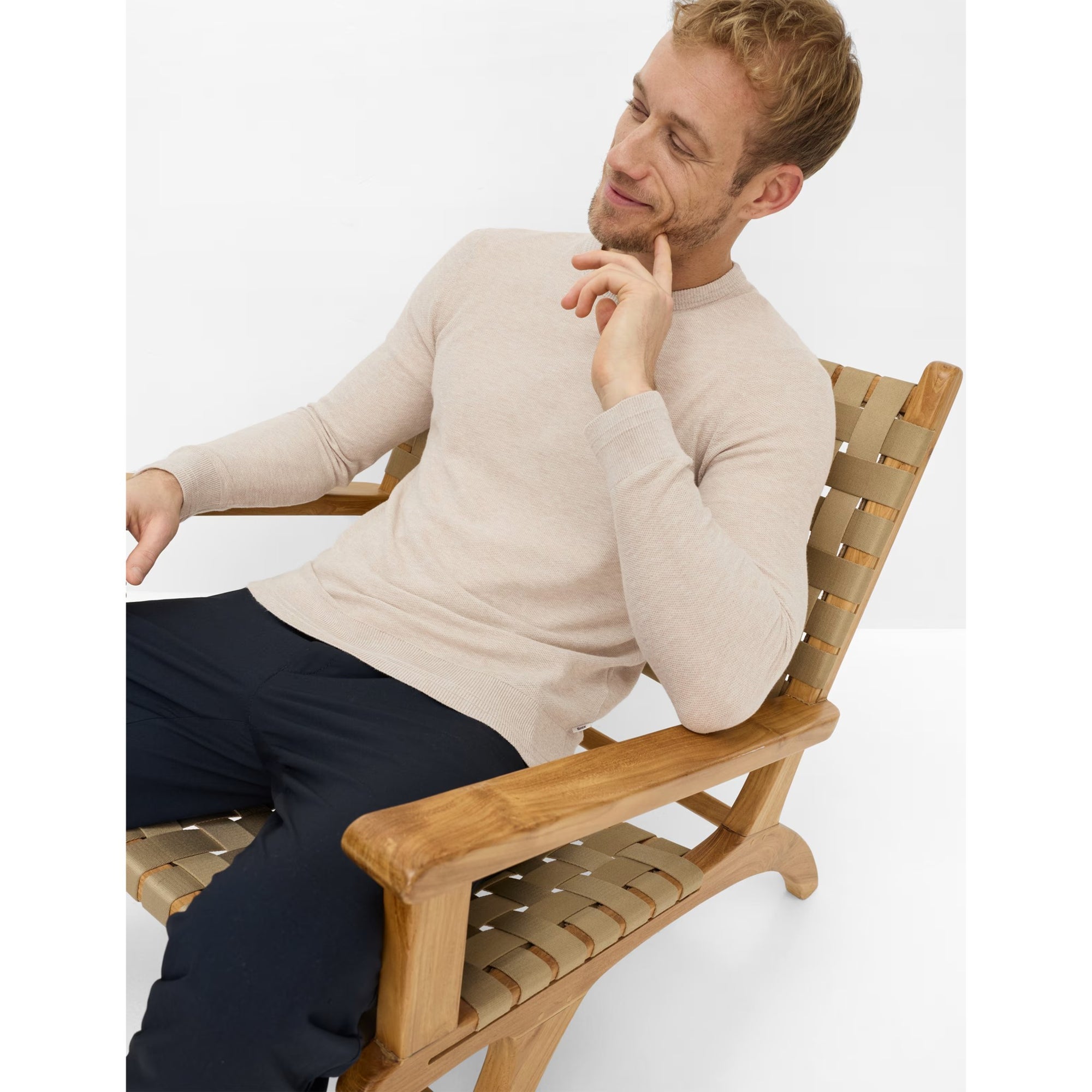 Man sitting on a wooden chair wearing a beige sweater and dark pants.