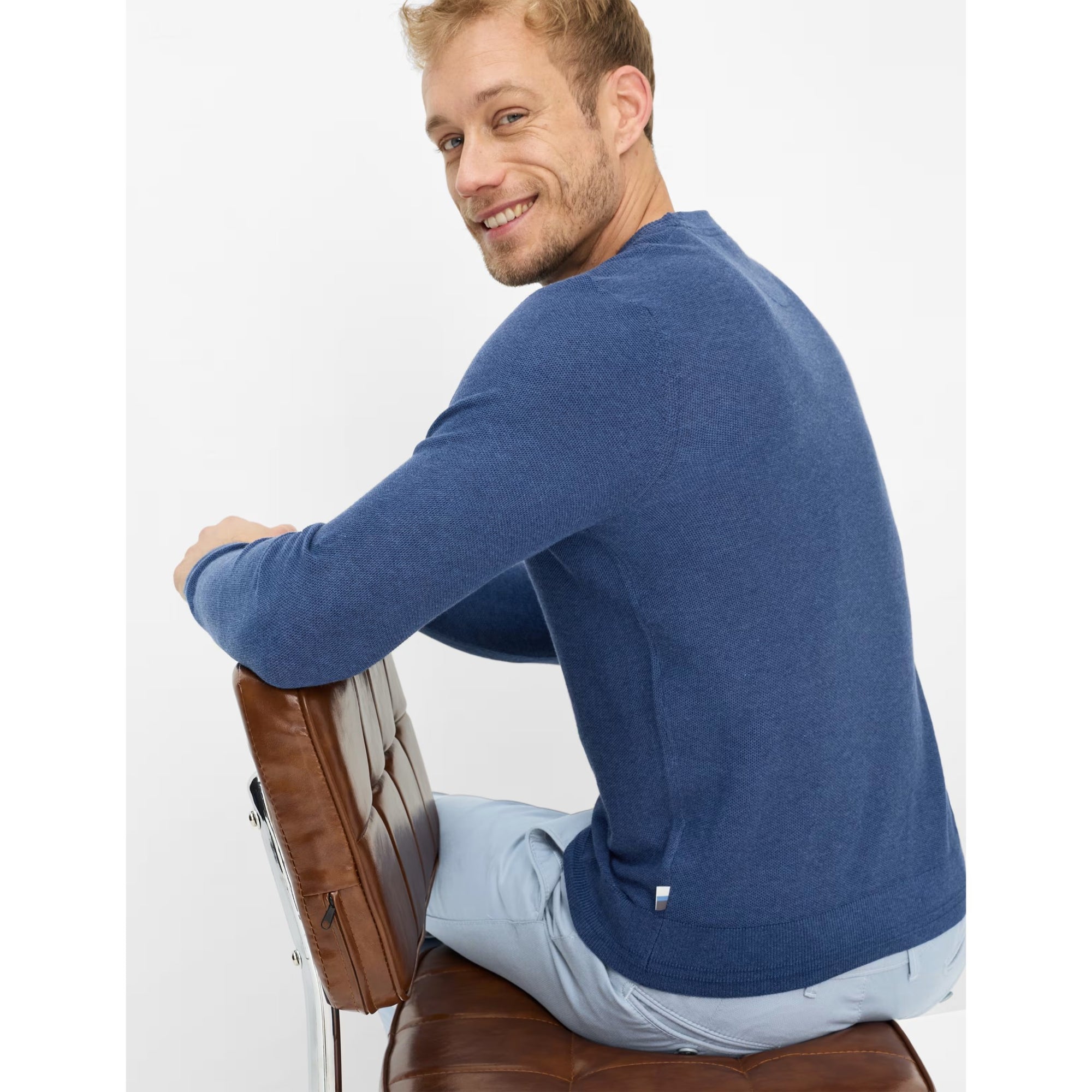 Man wearing a blue sweater sitting on a brown chair against a white background