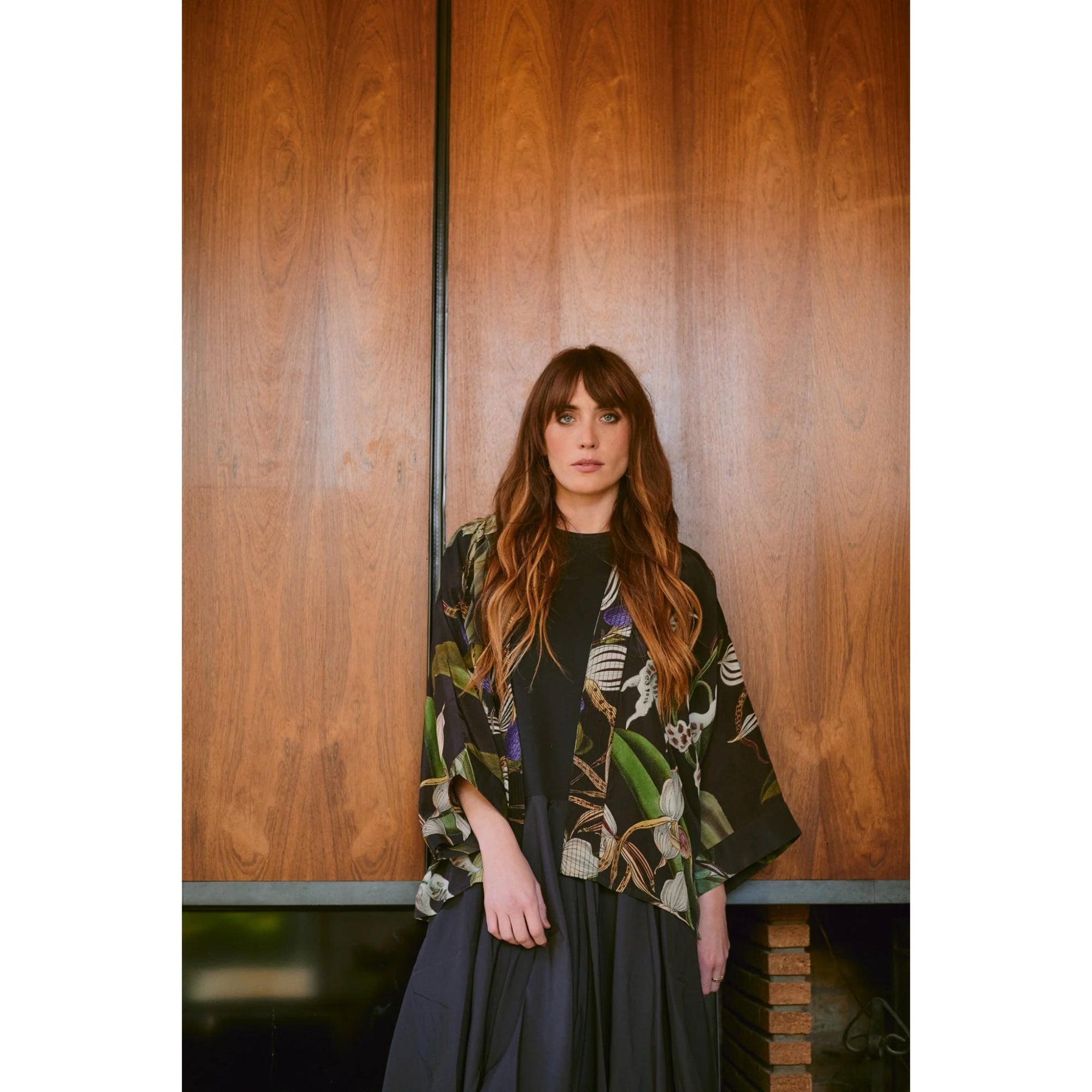 Woman wearing a floral kimono-style top against a wooden wall.