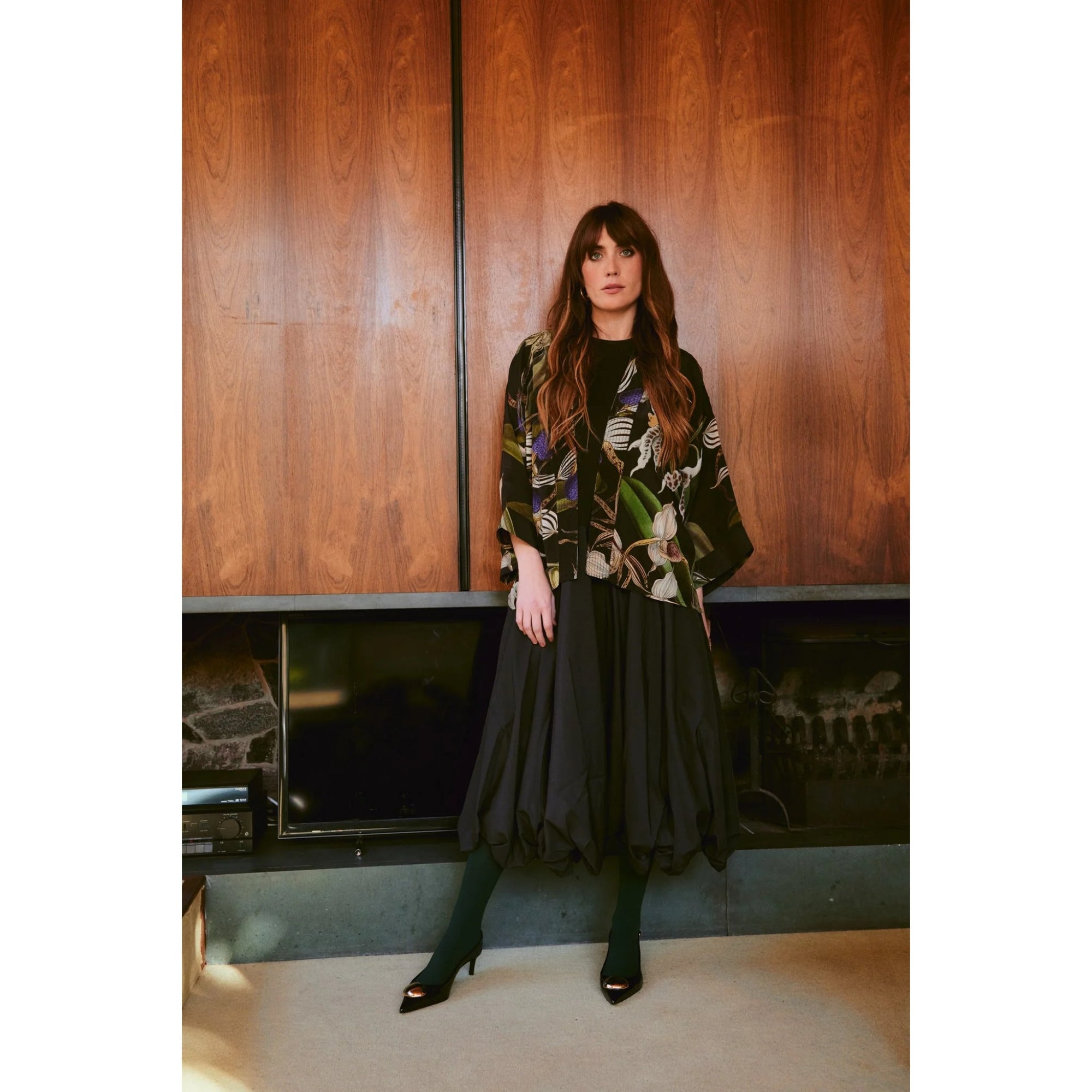 Woman in a floral jacket and black dress standing against a wooden paneled wall.