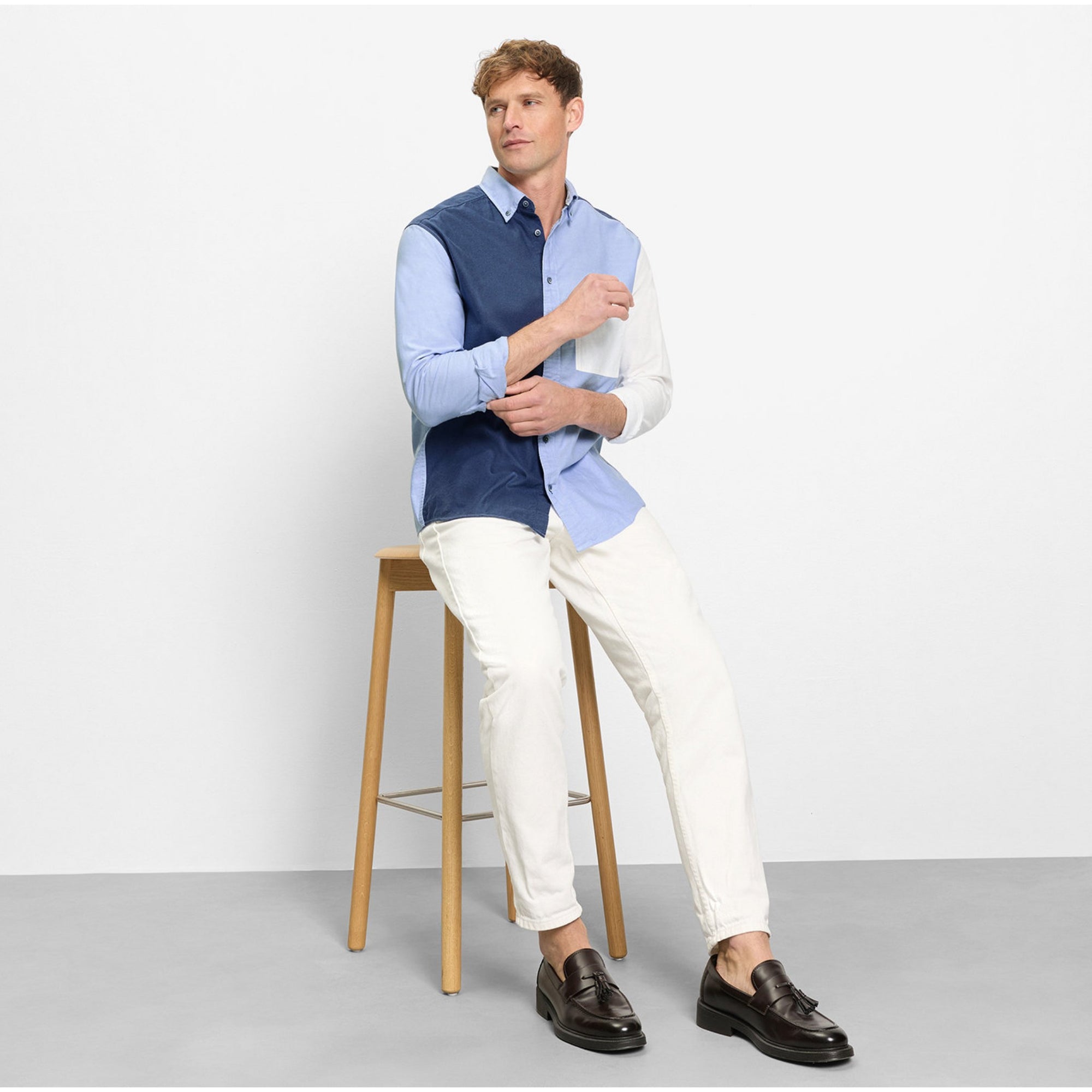 Man sitting on a stool wearing a blue vest over a light blue shirt, white pants, and brown shoes.