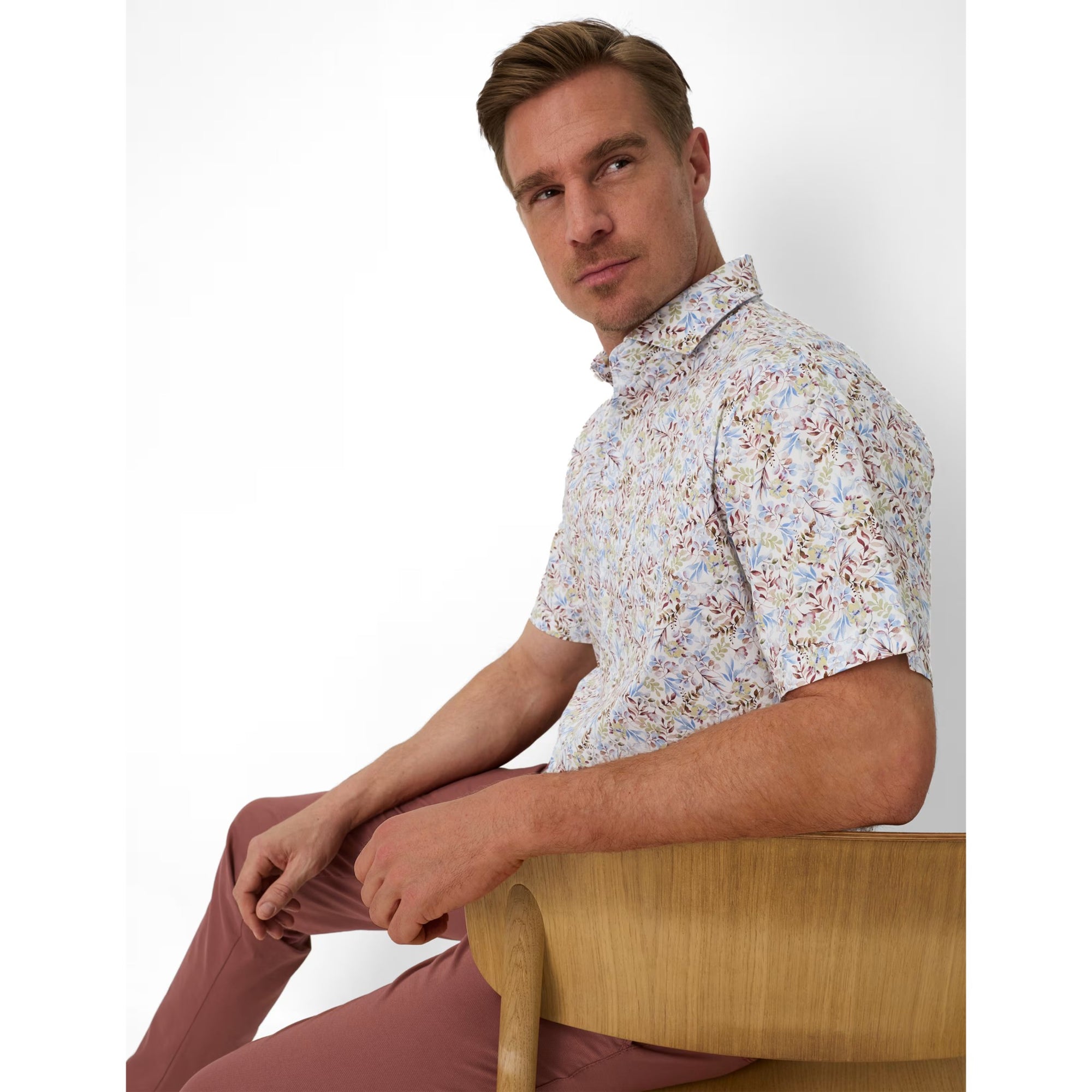 Man sitting on a chair wearing a patterned shirt and maroon pants against a white background