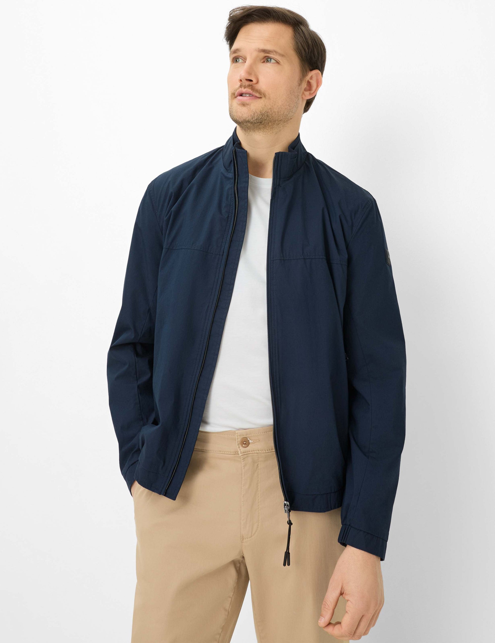 Man wearing a navy jacket over a white shirt and beige pants on a white background