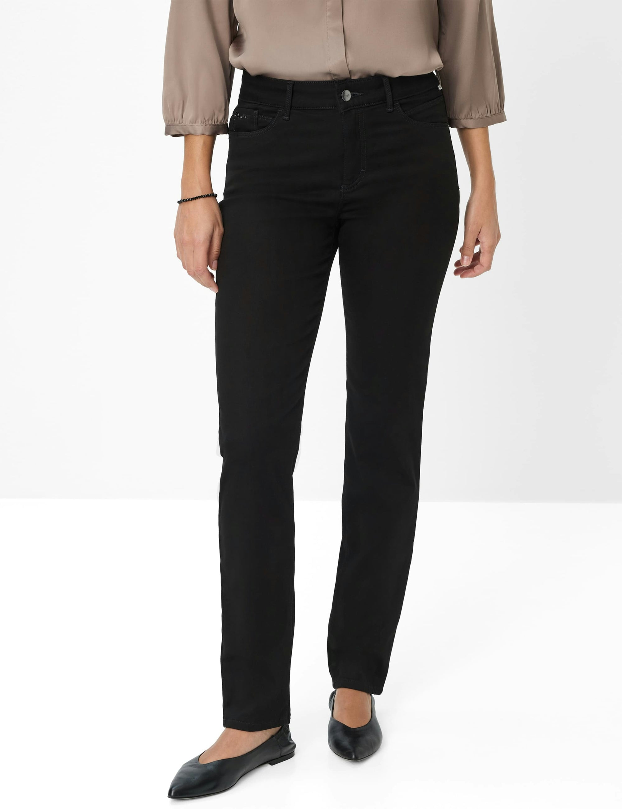 A model wearing black slim-fit jeans, paired with a taupe blouse and black flats, shown from the waist down against a white studio background.