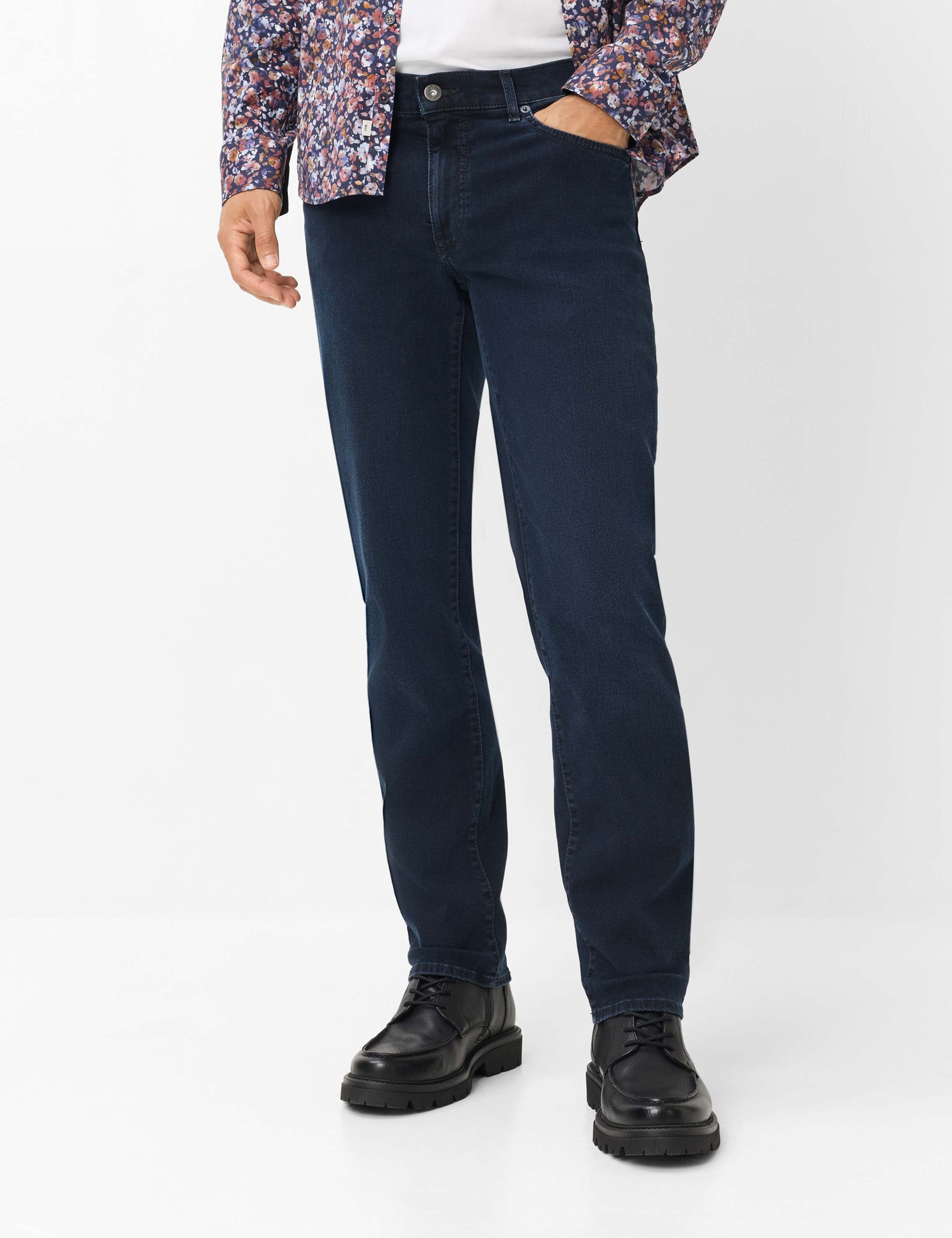 A person wearing dark navy blue Brax slim-fit jeans, styled with a patterned button-up shirt and black leather shoes, photographed against a clean white studio background.