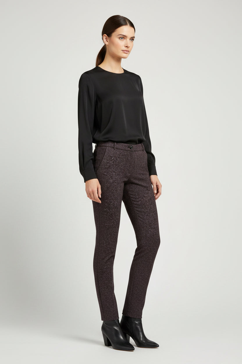 A woman modeling a black long-sleeve top and dark brown textured slim-fit trousers with black ankle boots, shot against a light gray studio background.