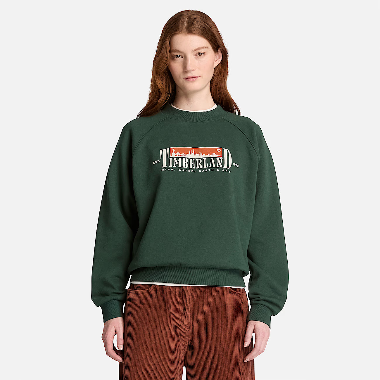 A model wearing a dark forest green Timberland crewneck sweatshirt with a 'TUMBLERLAND' graphic on the chest, paired with brown corduroy pants, against a light gray background.