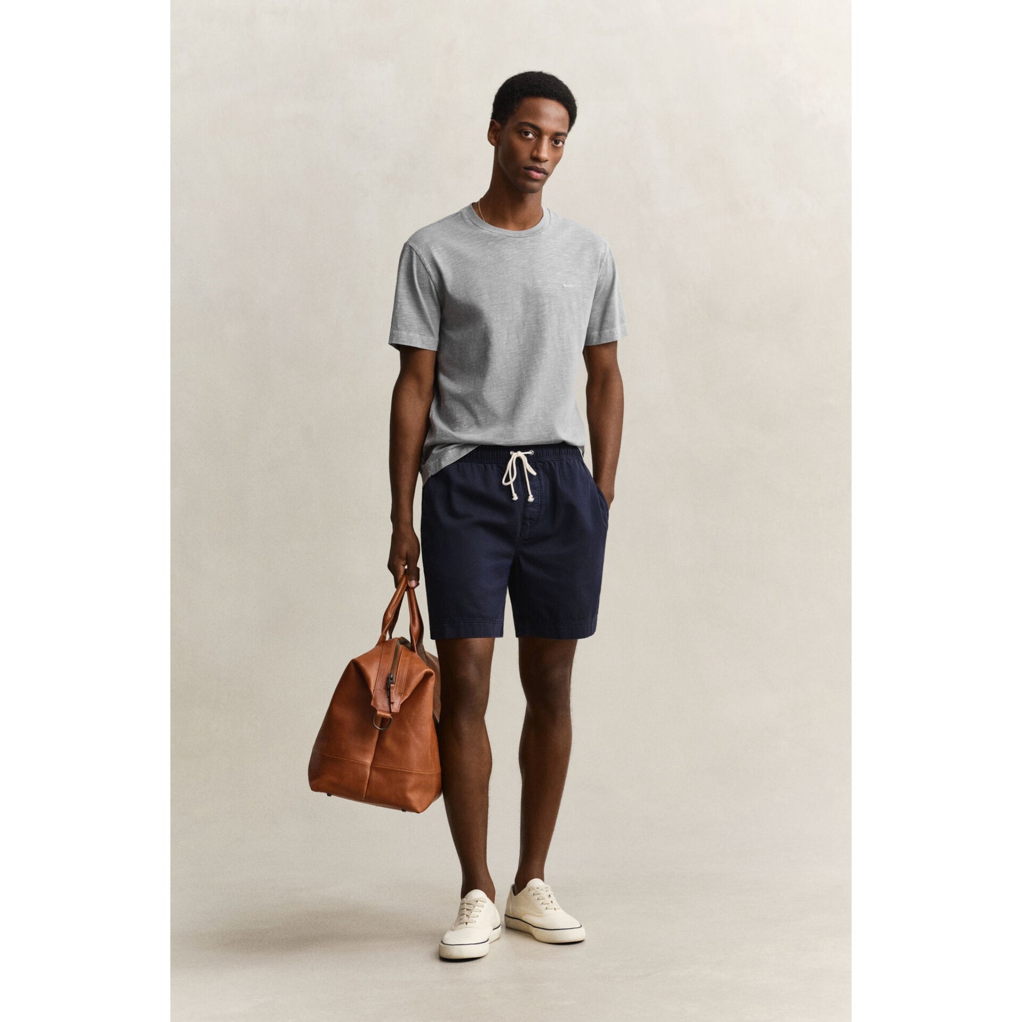 Man wearing a gray t-shirt and navy shorts, holding a brown leather bag on a beige background