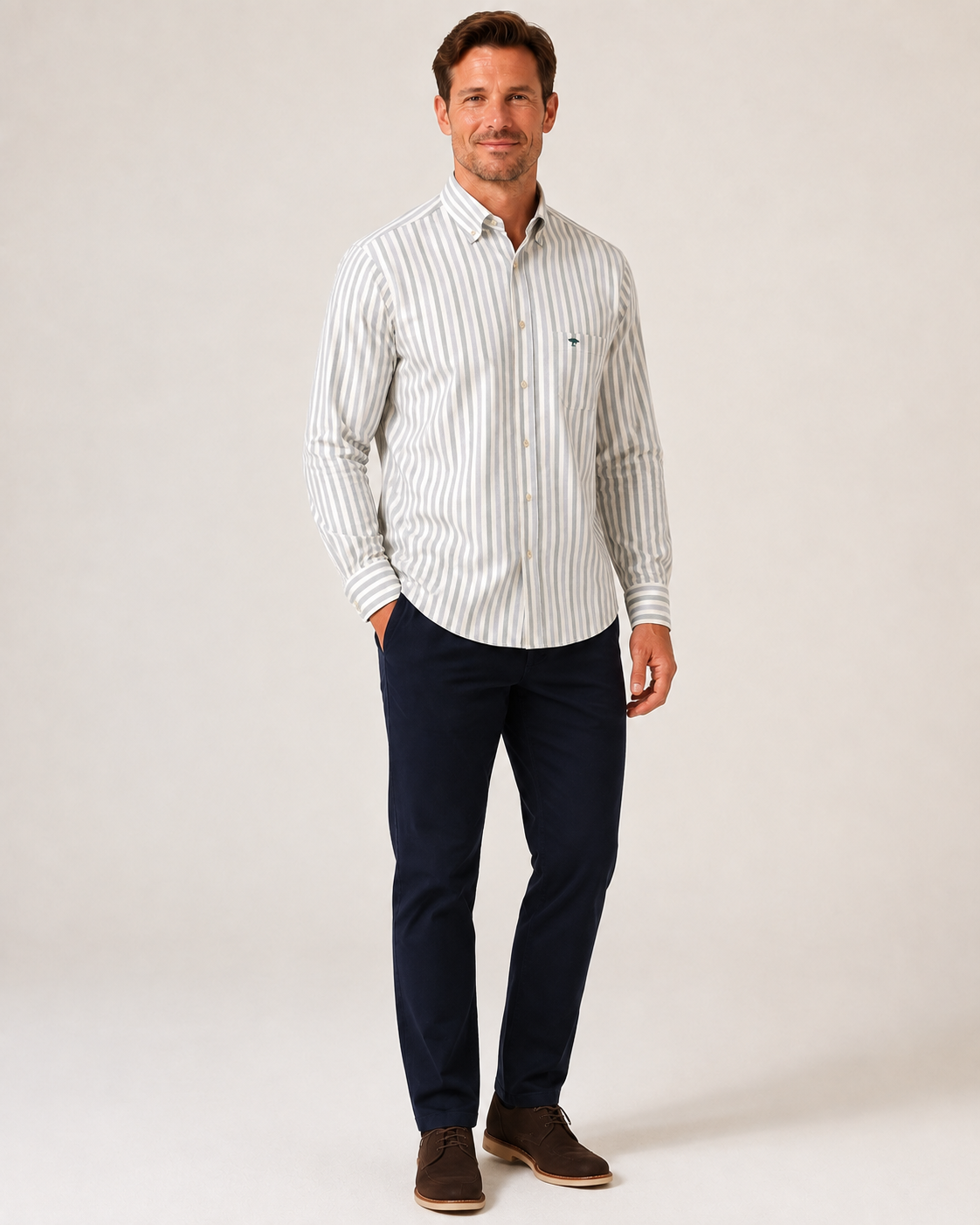 A man modeling a long-sleeve button-down shirt with thin vertical grey and pale teal stripes, paired with navy trousers and brown shoes, against a light beige studio background.