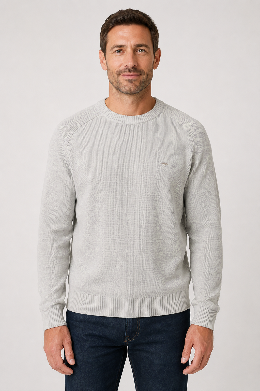 A man wearing a light gray crew-neck knit sweater and dark blue jeans, standing against a neutral light gray background.