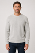 A man wearing a light gray crew-neck knit sweater and dark blue jeans, standing against a neutral light gray background.